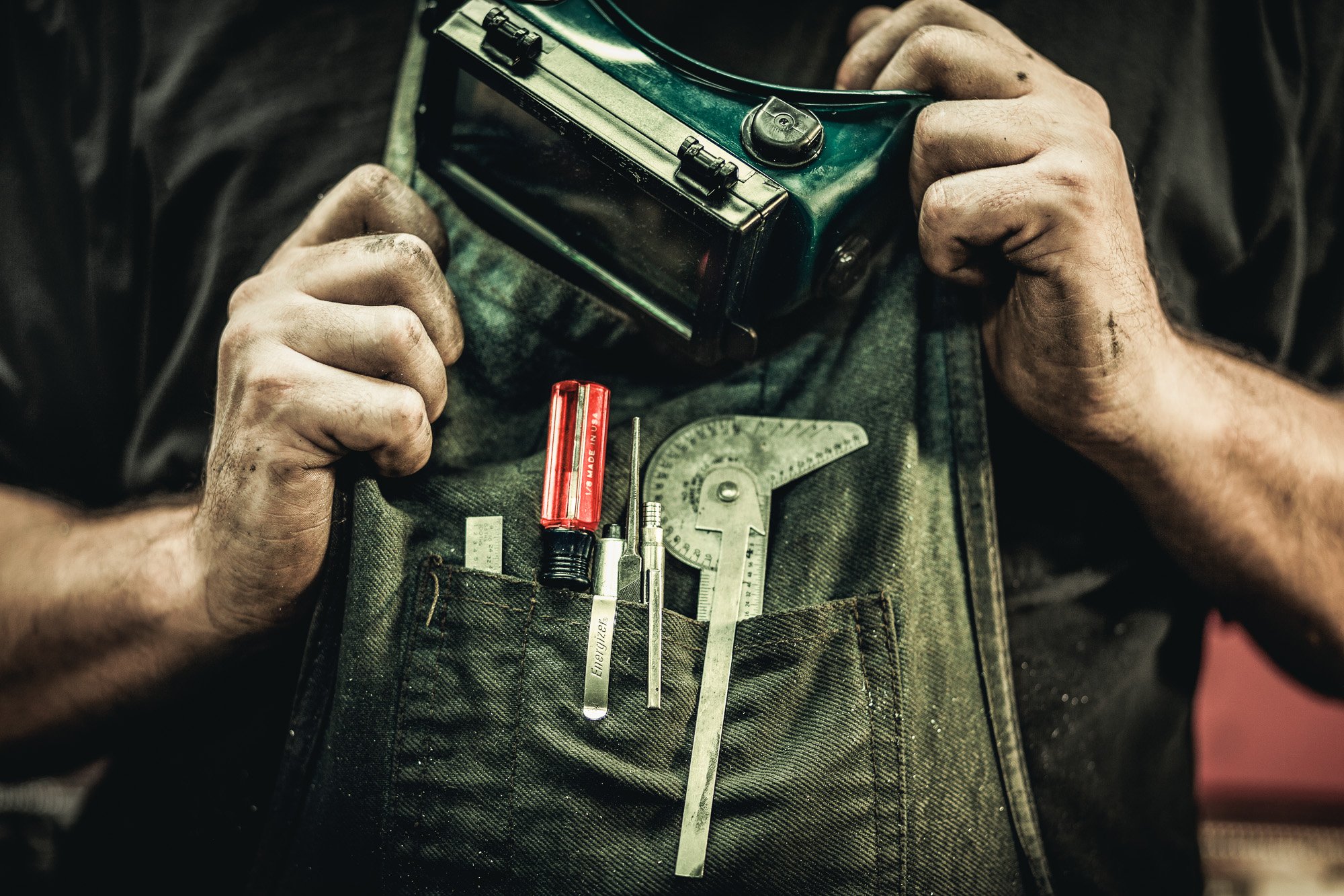 Close-up of a mechanic's dirt-covered hands holding a flashlight, with tools including a thermometer, screwdriver, and wrench in the shirt pocket, on the front of a green working apron.