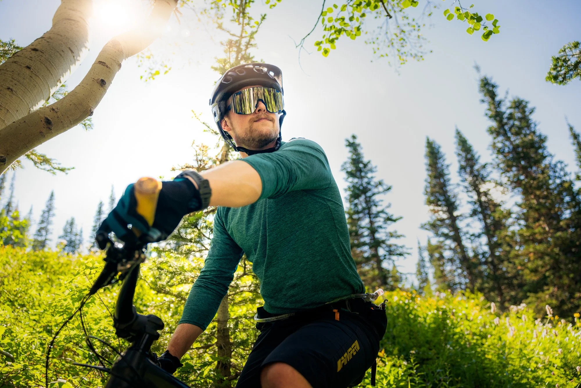 A person riding a mountain bike through a forest, wearing a helmet and reflective goggles, with sunlight filtering through trees.