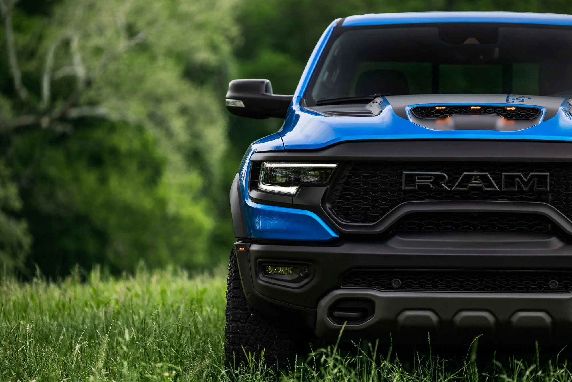 Close-up of the front part of a blue RAM pickup truck parked on grass with trees in the background.