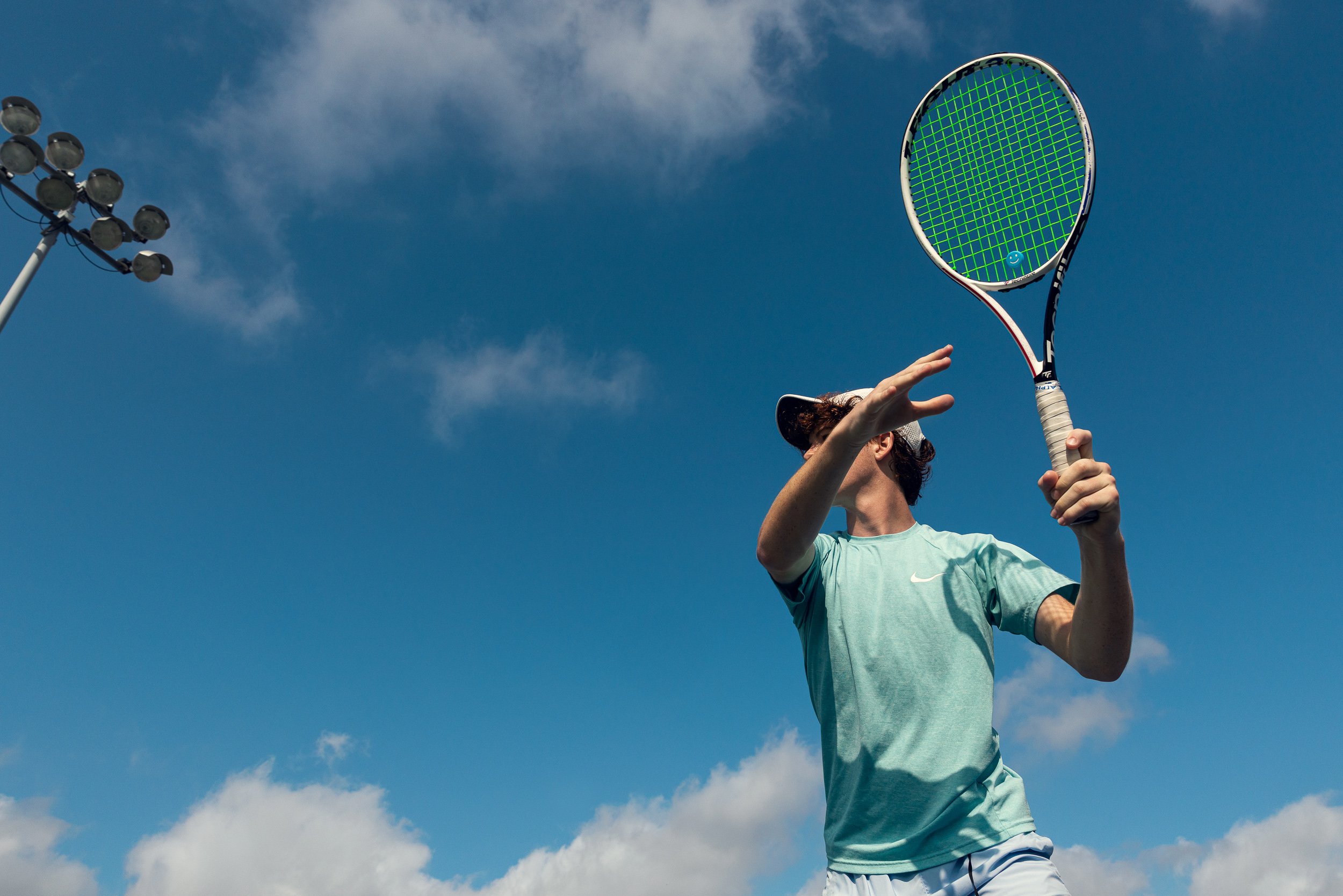Tennis player preparing to hit a shot on an outdoor court under a blue sky with some clouds, next to floodlights.