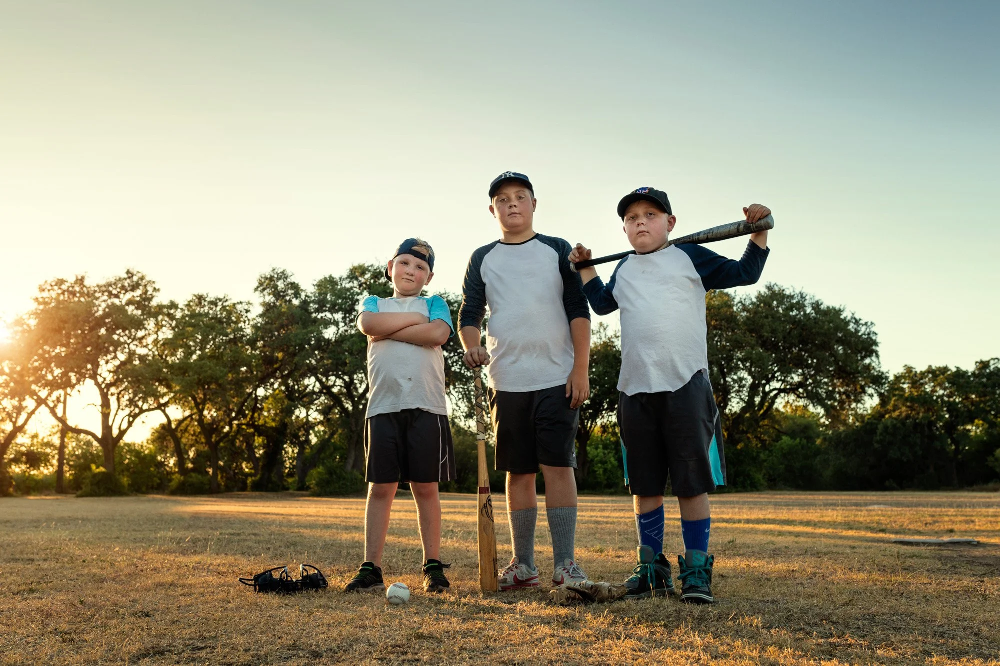 Three young boys in baseball uniforms standing on a grassy field at sunset, with trees in the background, holding baseball equipment and a bat.