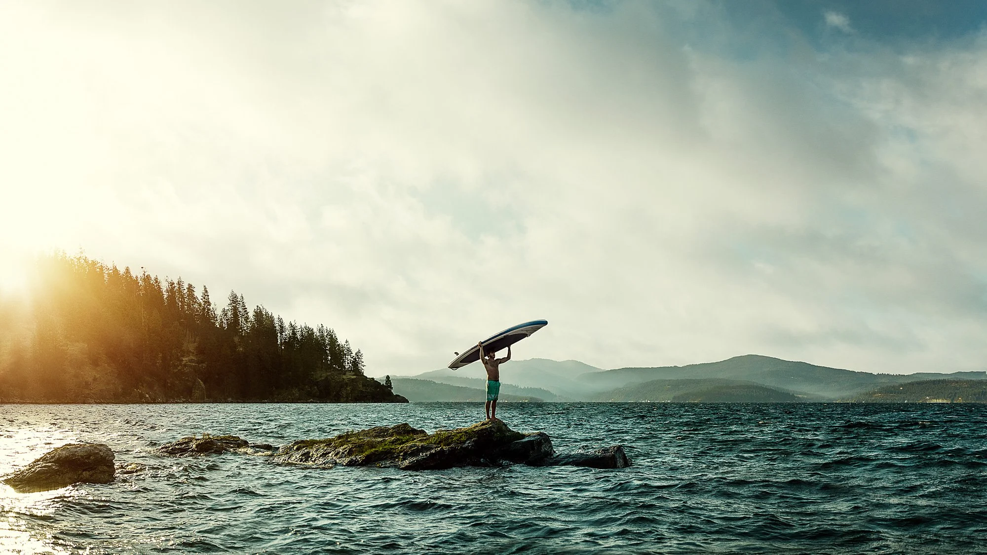 Person standing on a rock in the water, holding a surfboard above their head, with a scenic background of hills, trees, and a cloudy sky.