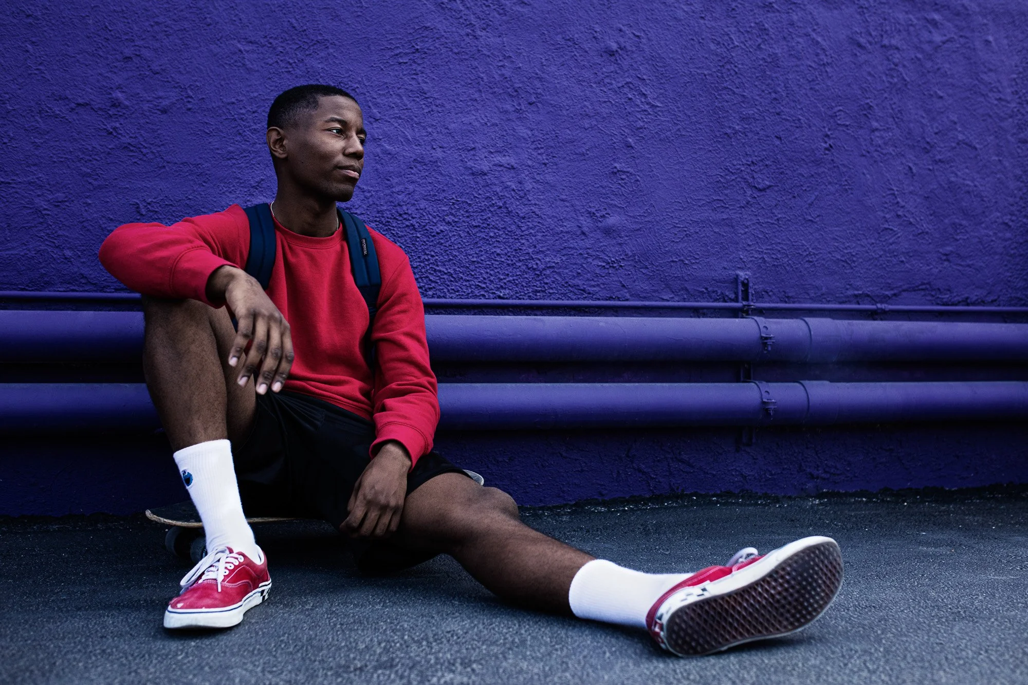 Young man in red sweatshirt and black shorts sitting on the ground with his back against a purple wall, wearing red sneakers and white socks, with a blue backpack, looking contemplative.