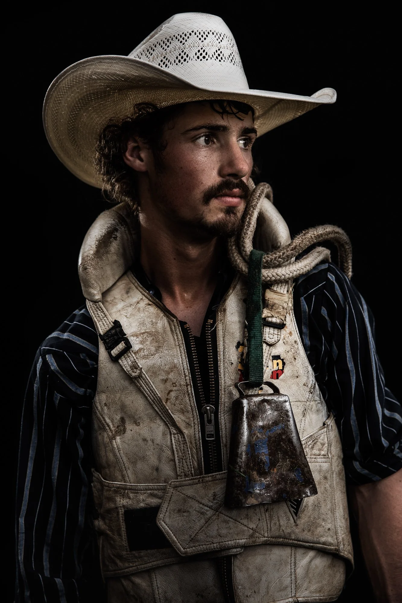 A man wearing a cowboy hat, striped shirt, and a dirty vest with a large metal object hanging around his neck, set against a black background.