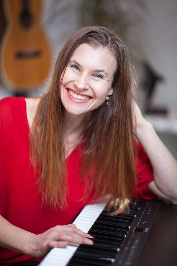 Anita Stryker, speech-language pathologist in Portland, Oregon. A woman with long brown hair and a red top sitting at a piano and smiling at the camera.