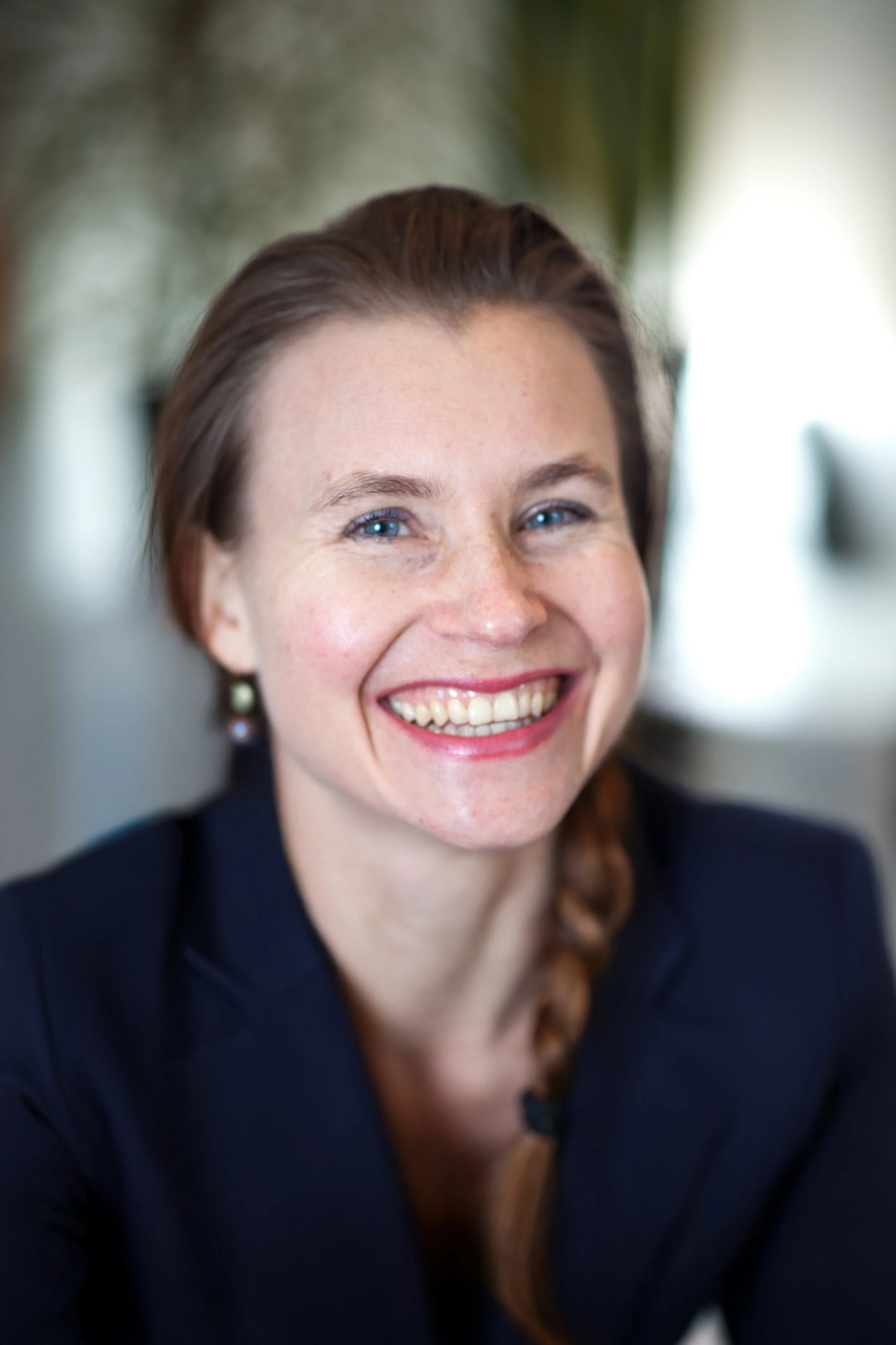 Anita Stryker, speech-language pathologist in Portland, Oregon. Close-up of a woman smiling with blue eyes, brown hair in a braid, wearing a dark blazer, and earrings, with a blurred indoor background.