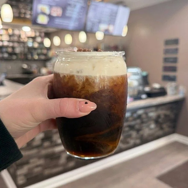 A hand holding up a glass cup of coffee with cold foam on top. Set inside West View Brew coffee shop.