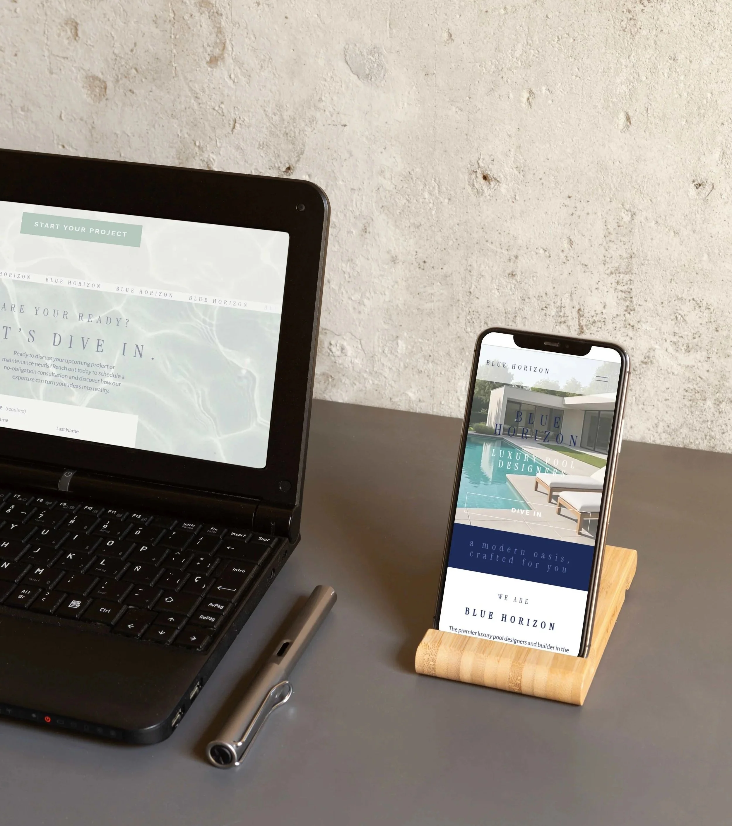 A laptop and a smartphone displaying the website design of Blue Horizon Pools by Hailey Osborne Designs, placed on a gray desk against a textured white wall.
