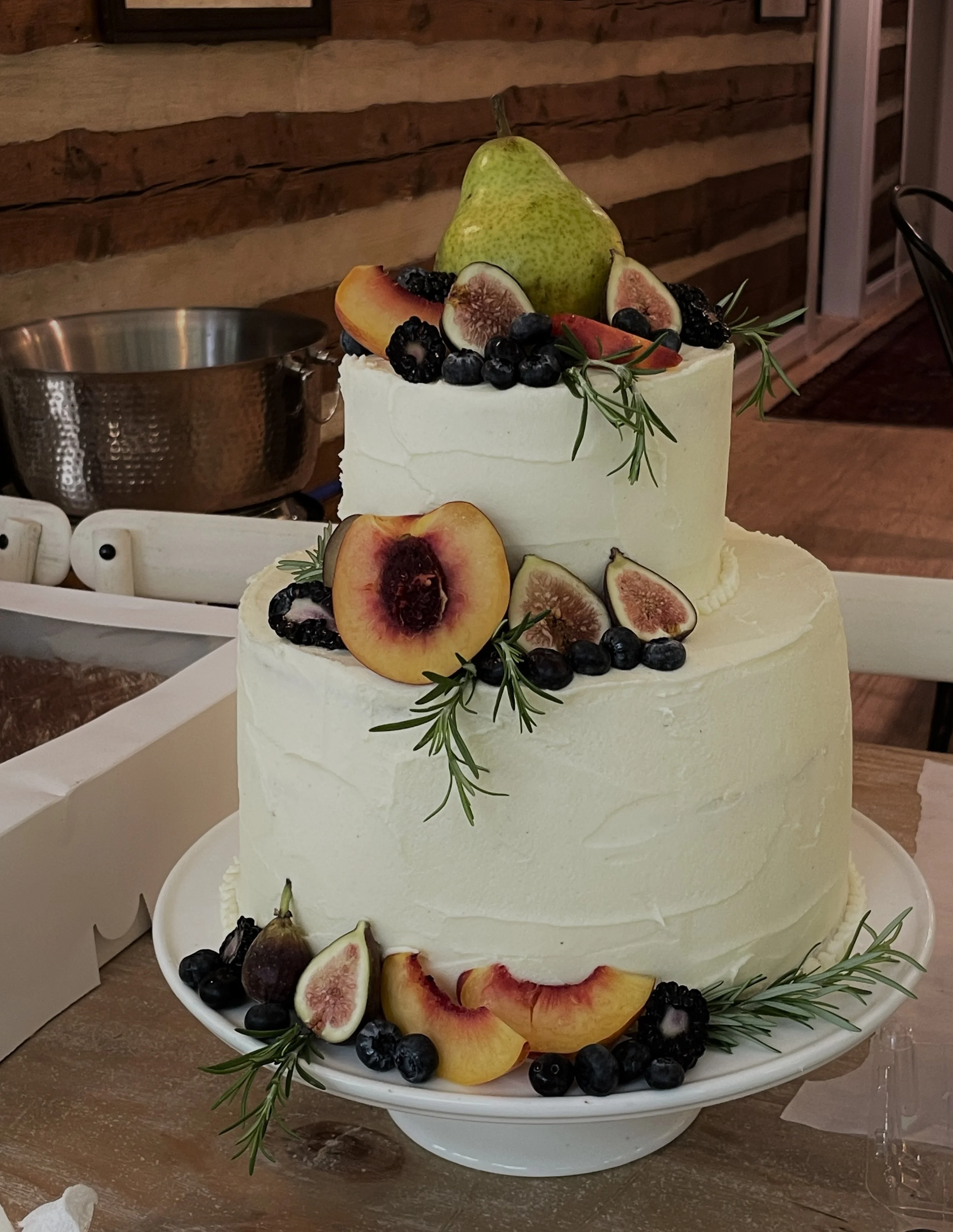 Two-tiered white frosted cake decorated with fresh figs, peaches, blueberries, blackberries, and sprigs of rosemary. Hailey Osborne Designs.