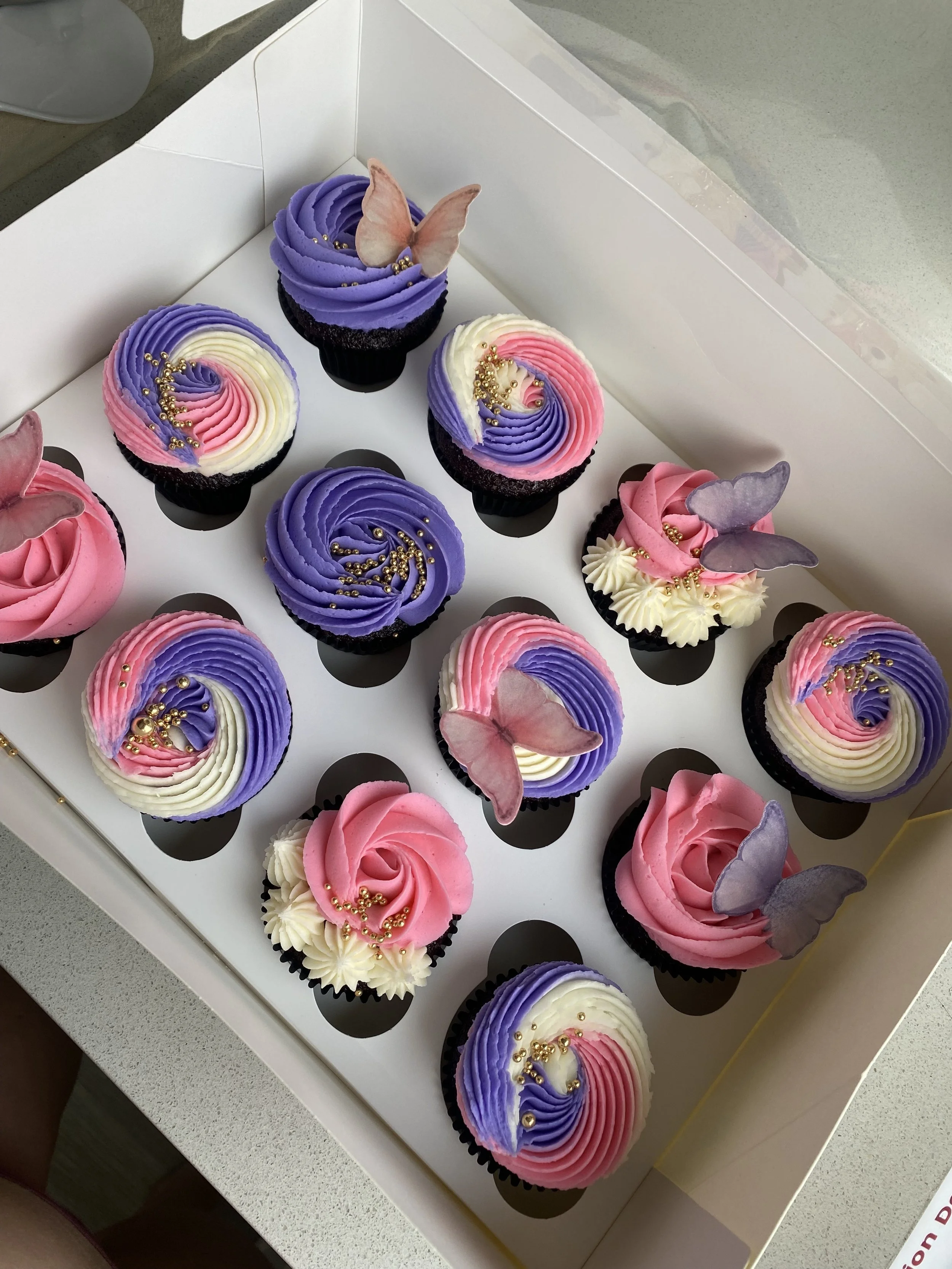 A box of decorated cupcakes with pink, purple, and white frosting, each adorned with edible butterflies and gold-colored edible beads.