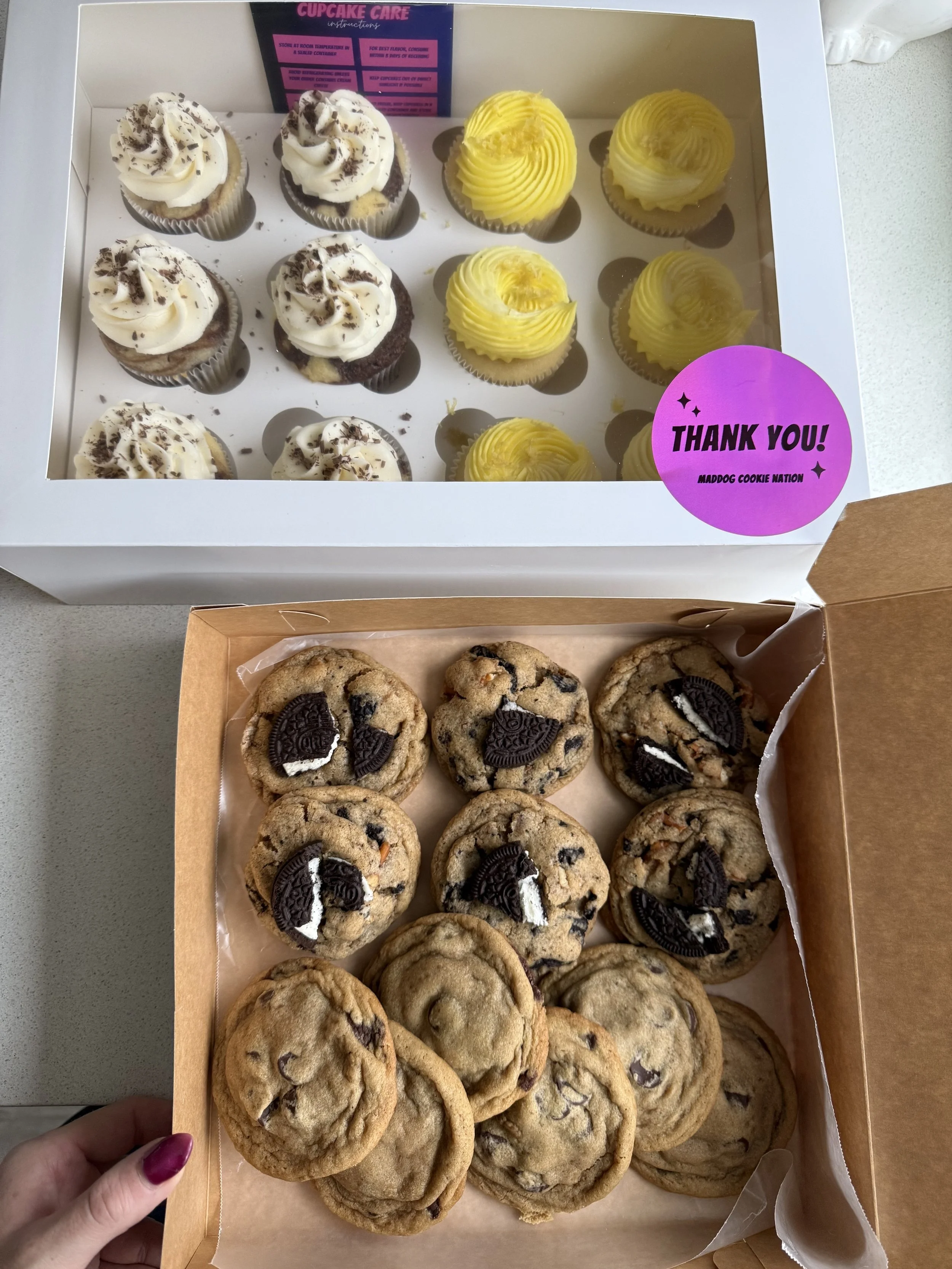 A box of cupcakes with yellow frosting and white cupcakes with chocolate sprinkles next to a box of chocolate chip cookies topped with Oreo cookies.