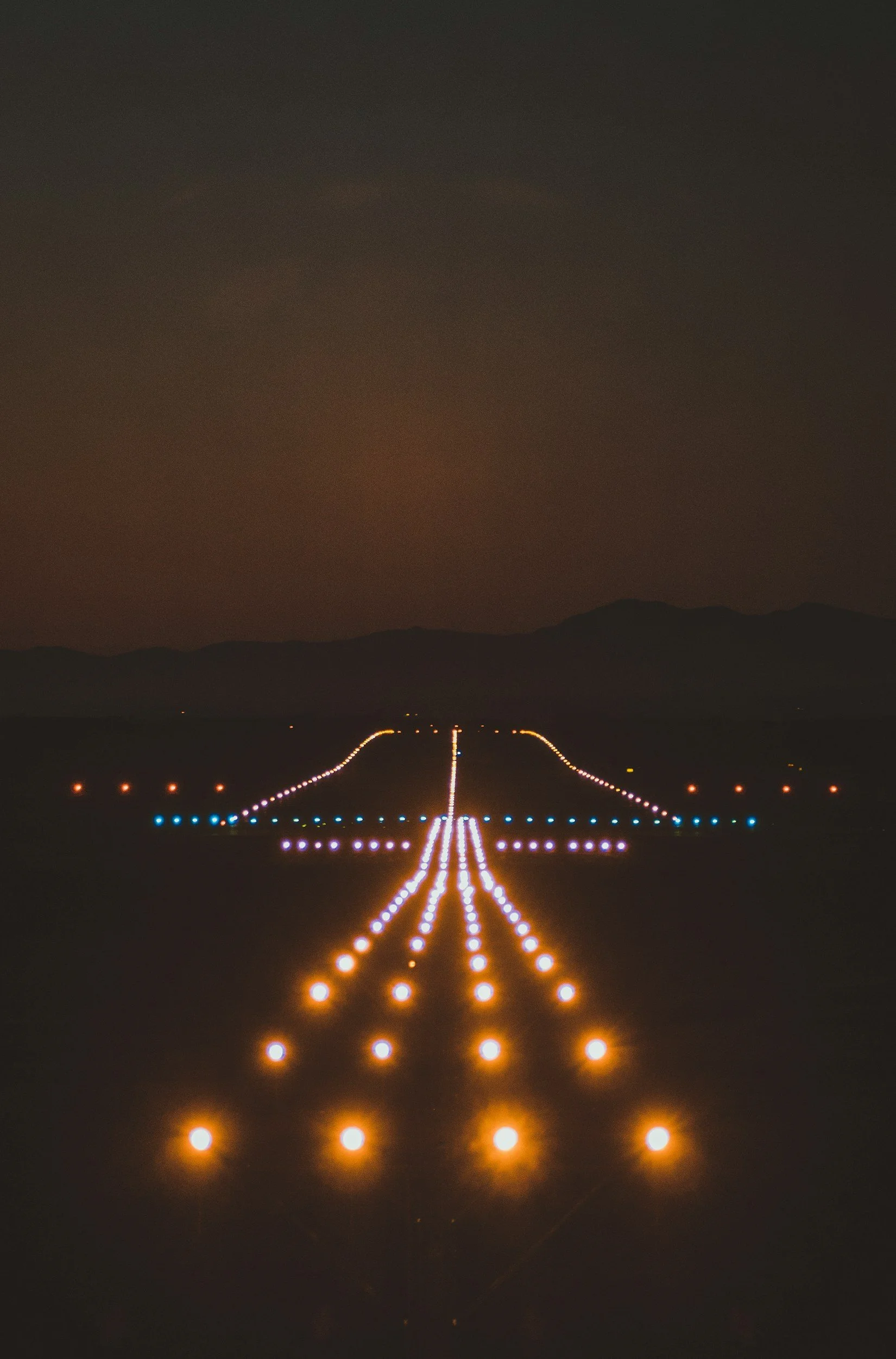 Nighttime view of an illuminated runway at an airport with colorful runway lights guiding the way, mountains visible in the background.