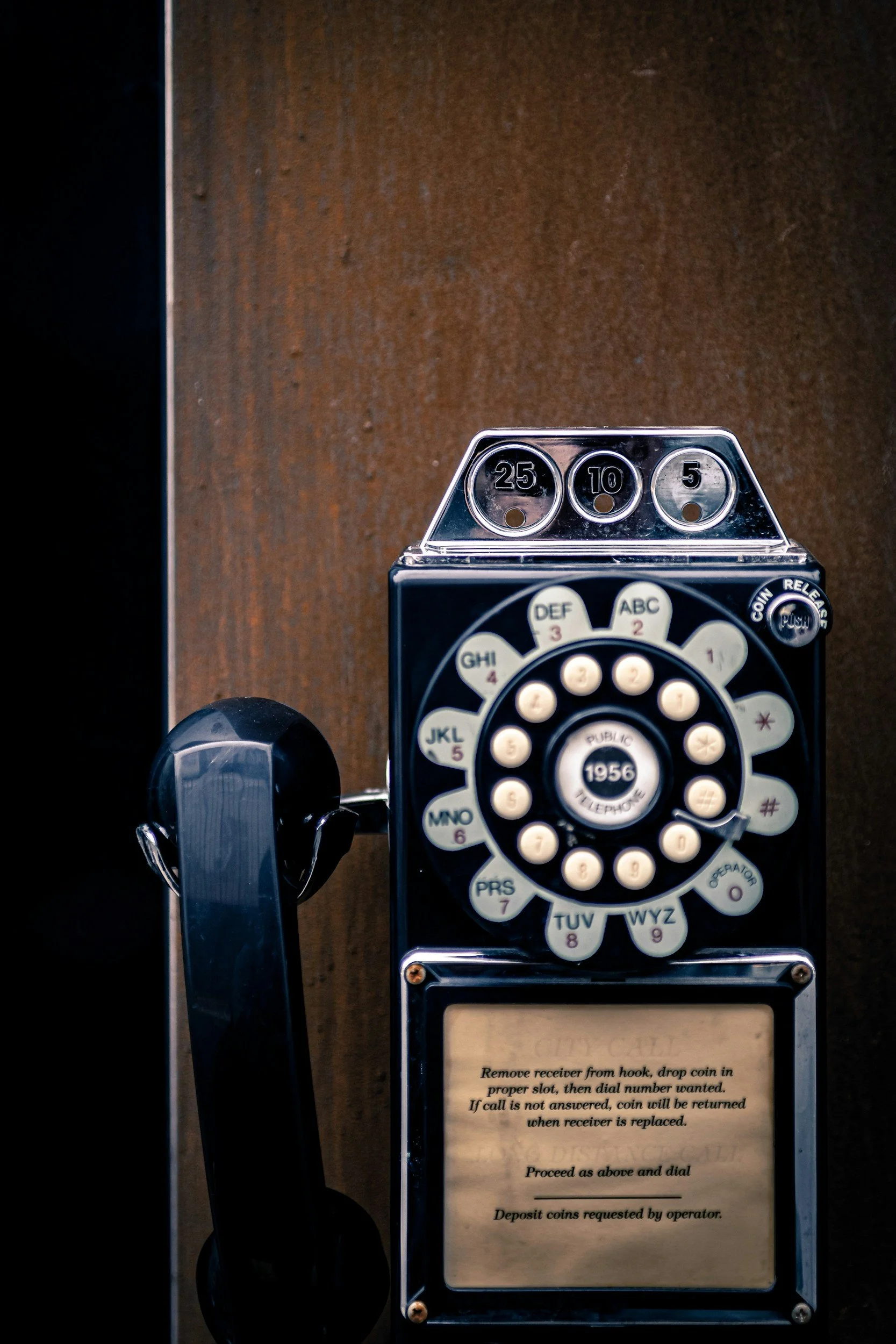 Vintage payphone with black handset, rotary dial, and instructions on a paper attached to the lower part of the phone, set against a brown background.