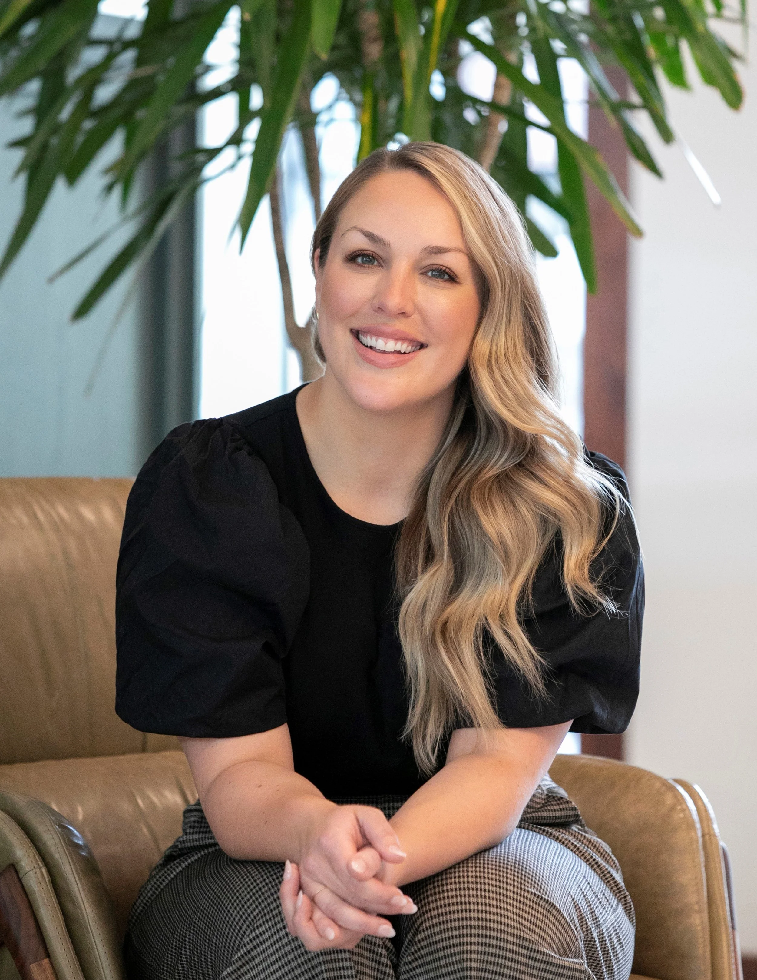 A woman with long wavy blonde hair smiling, sitting on a tan leather chair in front of a large green plant in an office or lounge area.