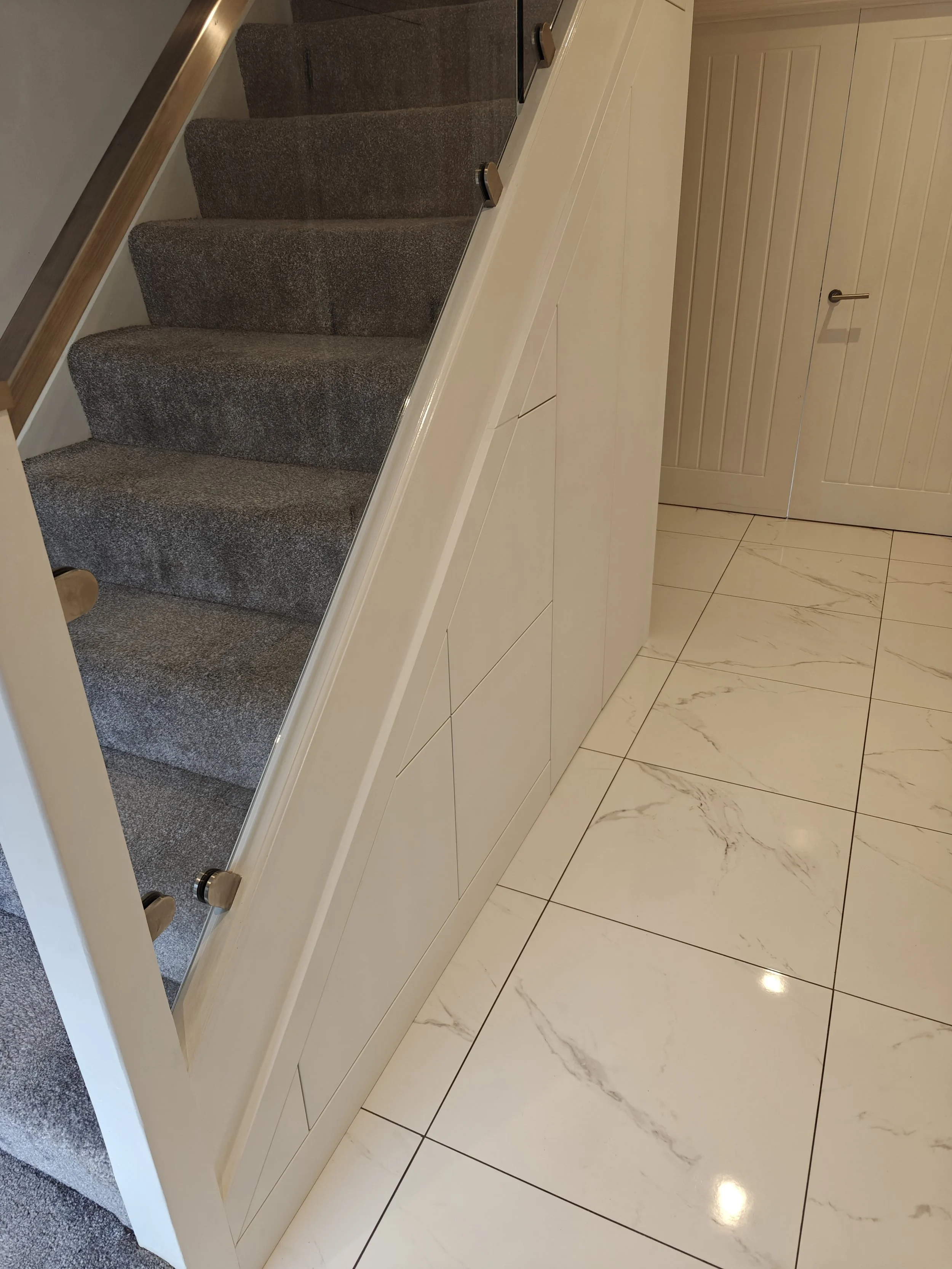 Interior view of a staircase with gray carpeted steps, white cabinetry or wall paneling along the side, and cream-colored marble tile flooring with subtle veining. There is a closed double door with vertical paneling and a handle in the background.