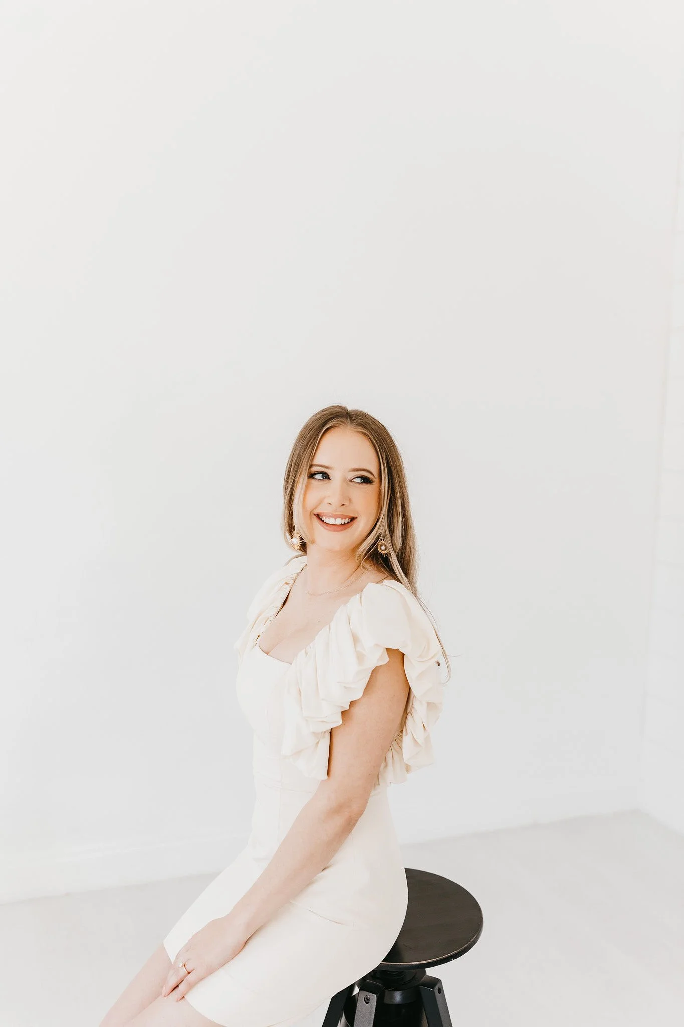A woman with long light brown hair wearing a cream-colored dress with ruffled sleeves, smiling while sitting on a black stool in a white room.