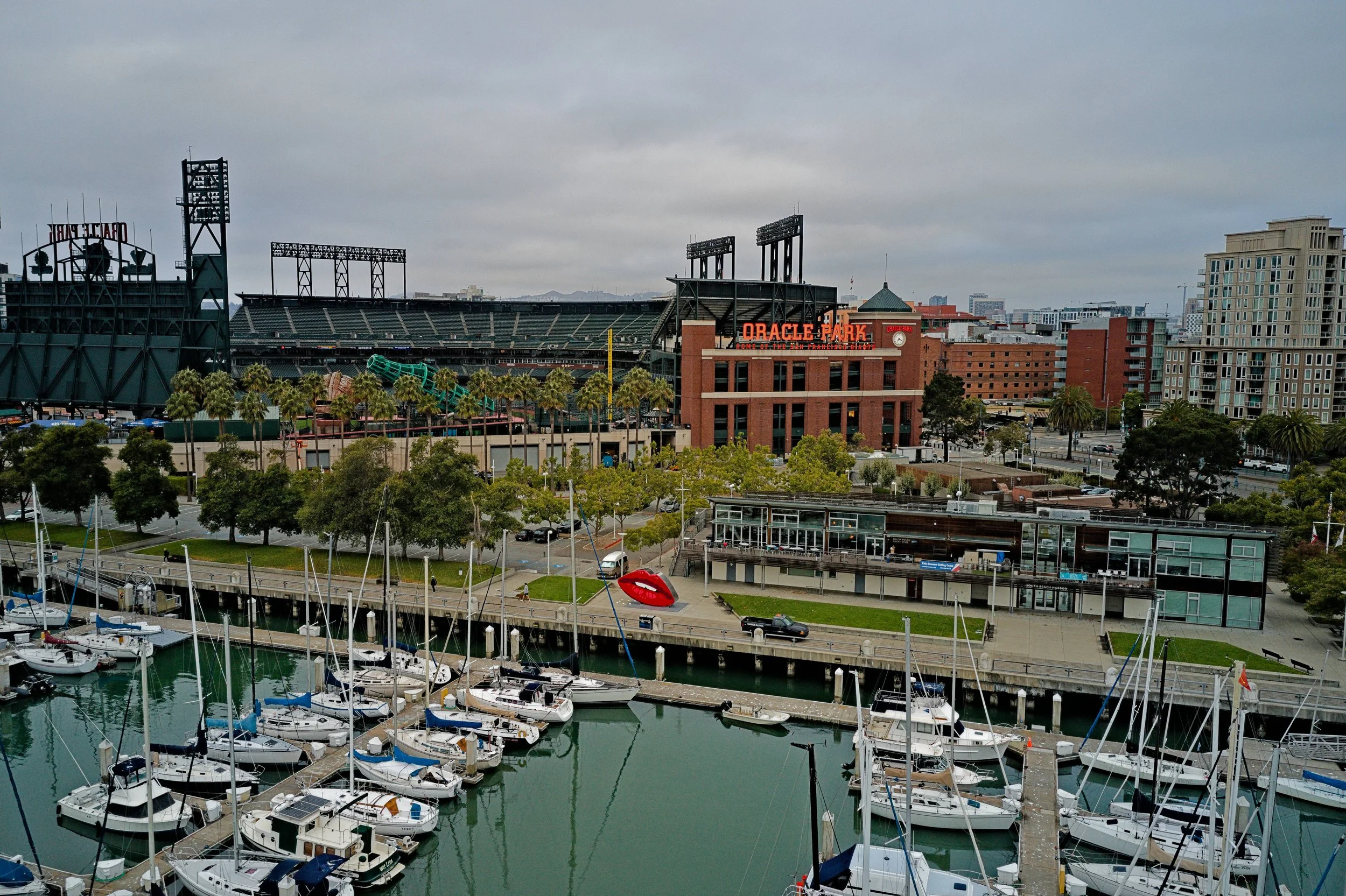 A marina with numerous sailboats docked, in front of a city skyline with tall buildings, including Oracle Park stadium with its sign and floodlights, under a gray cloudy sky.