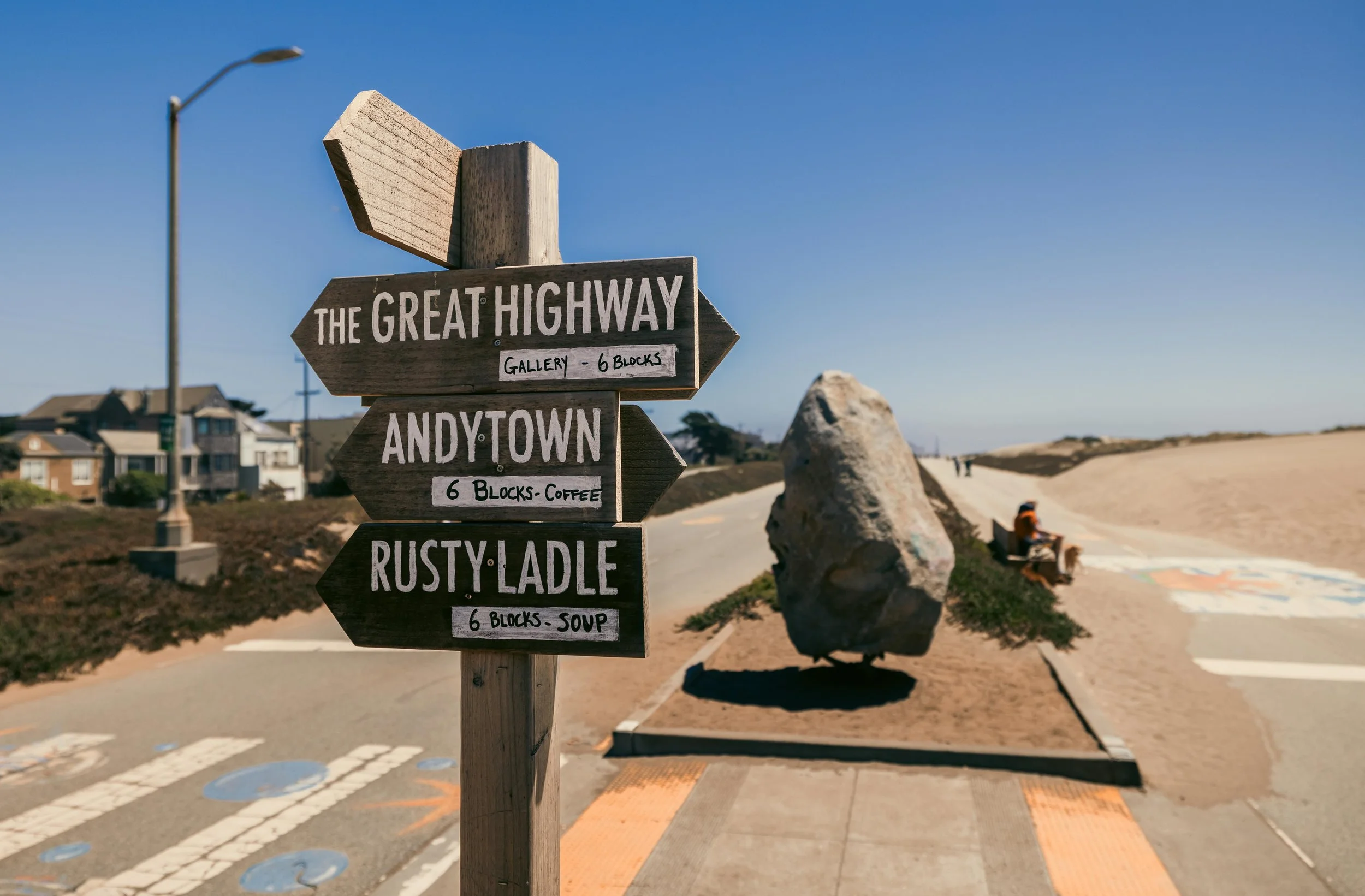 Wooden signpost on a sidewalk pointing to The Great Highway and nearby locations, with houses and a beach in the background under a clear blue sky.