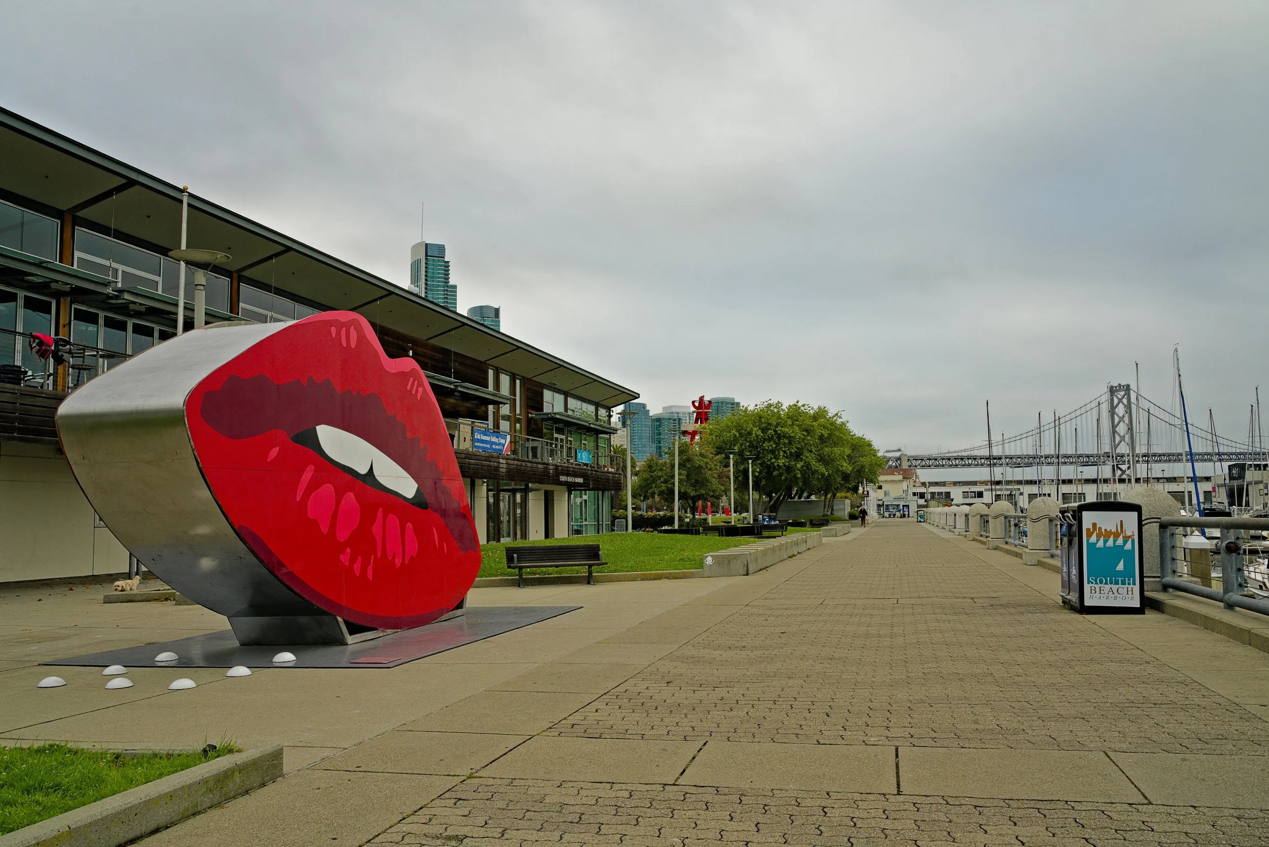 A large outdoor sculpture of red lips with white teeth, placed on a sidewalk near a building with glass windows and balconies. Background shows a waterfront, trees, and a view of a bridge and city skyline under a cloudy sky.