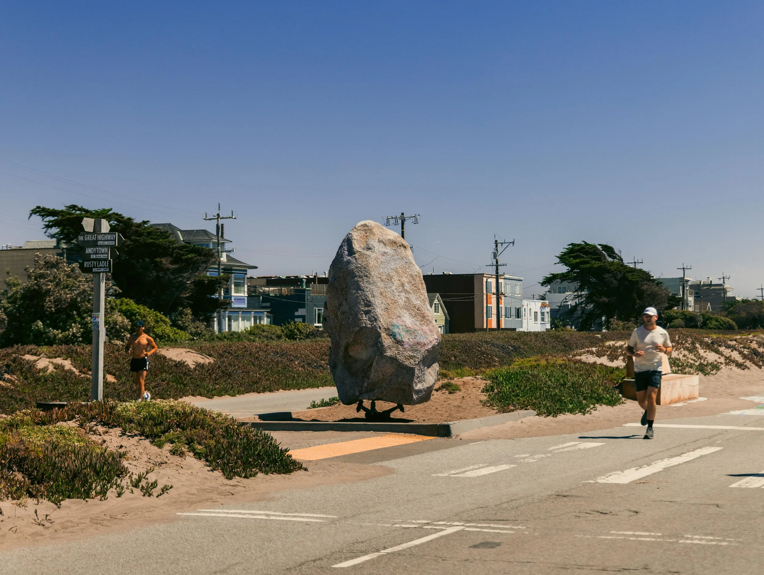 A large rock sculpture resembling a bird stands in a park, surrounded by trees, power lines, and residential buildings in the background. Two people are walking nearby, one shirtless man and one woman wearing a white shirt and black shorts.