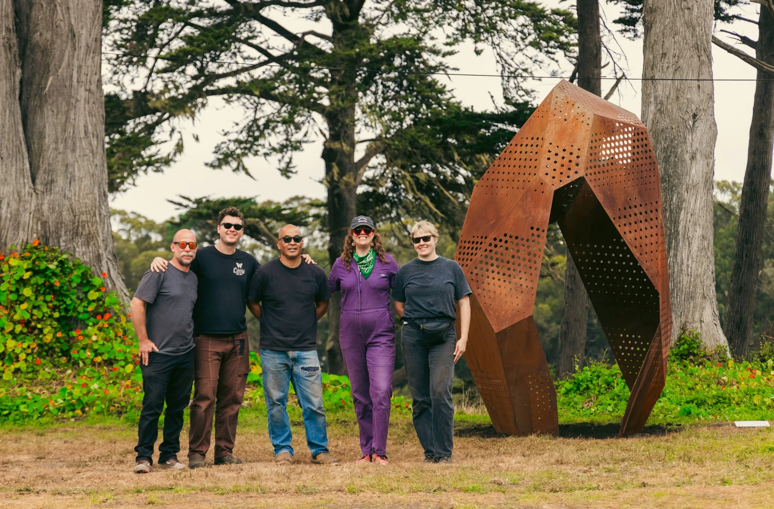 A group of five people standing outdoors in front of a large metal sculpture, surrounded by trees and greenery.