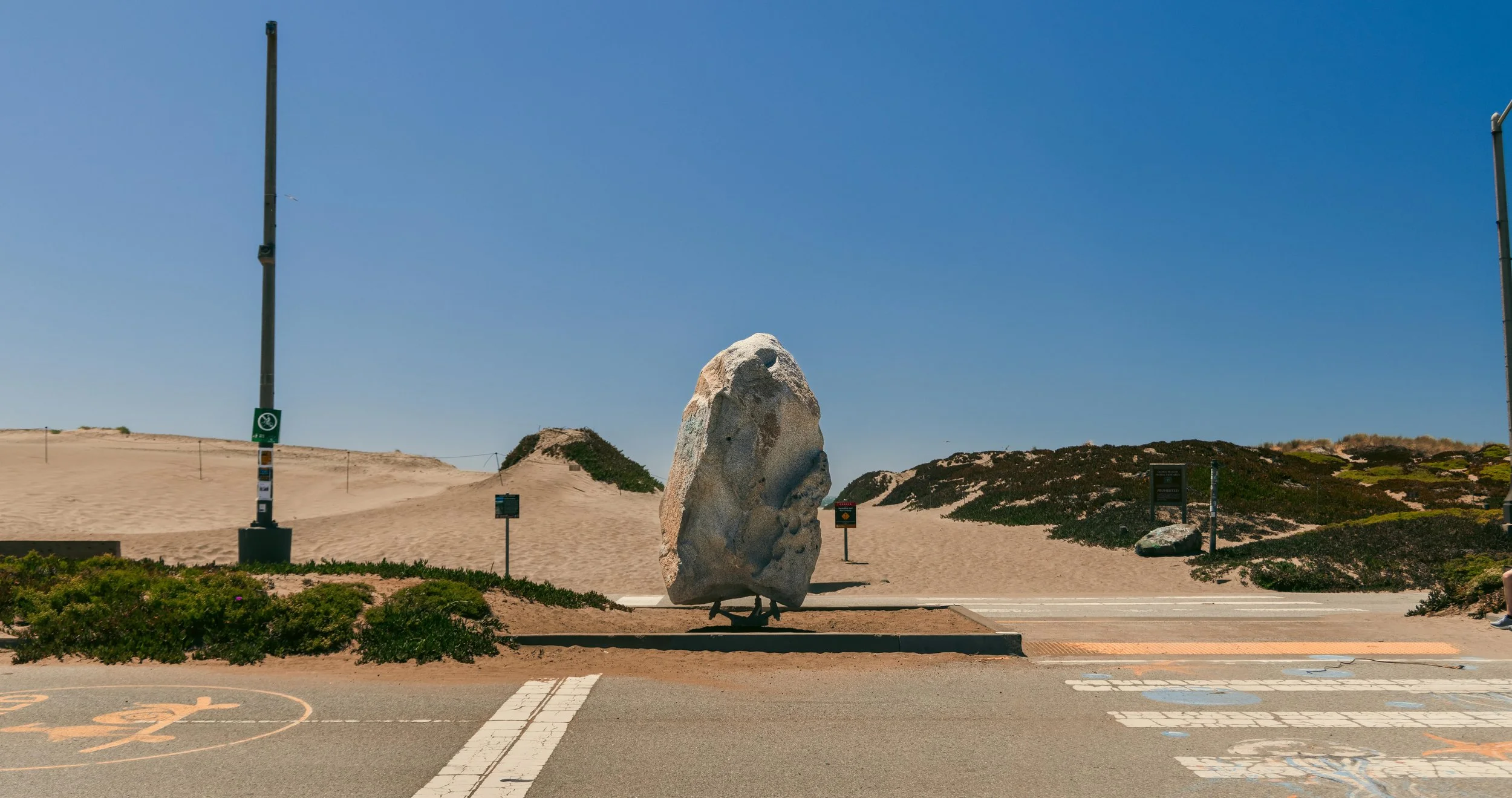 A large rock sculpture on a sidewalk in a sandy landscape with hills and sparse vegetation under a clear blue sky.