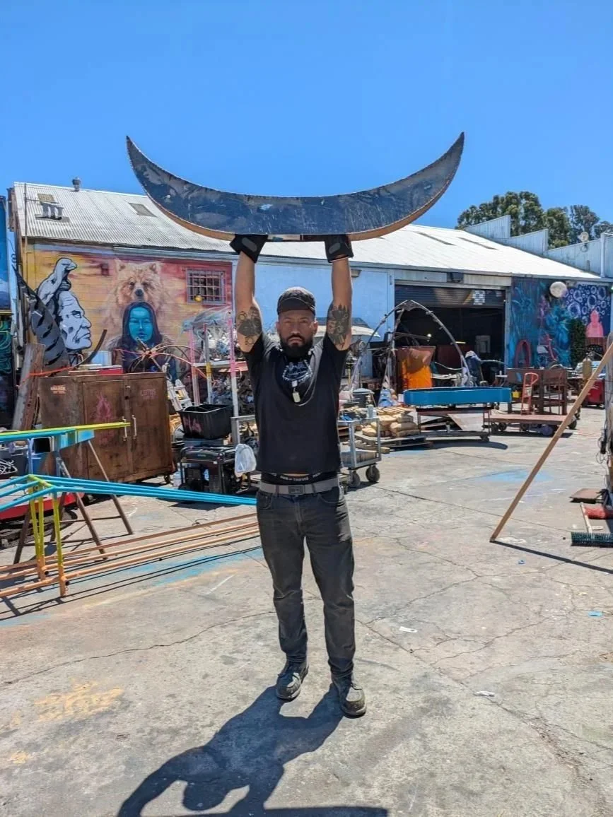 Artist Zulu Heru holding a large curved metallic sculpture above his head outdoors in a colorful, graffiti-covered area with various furniture and art supplies.