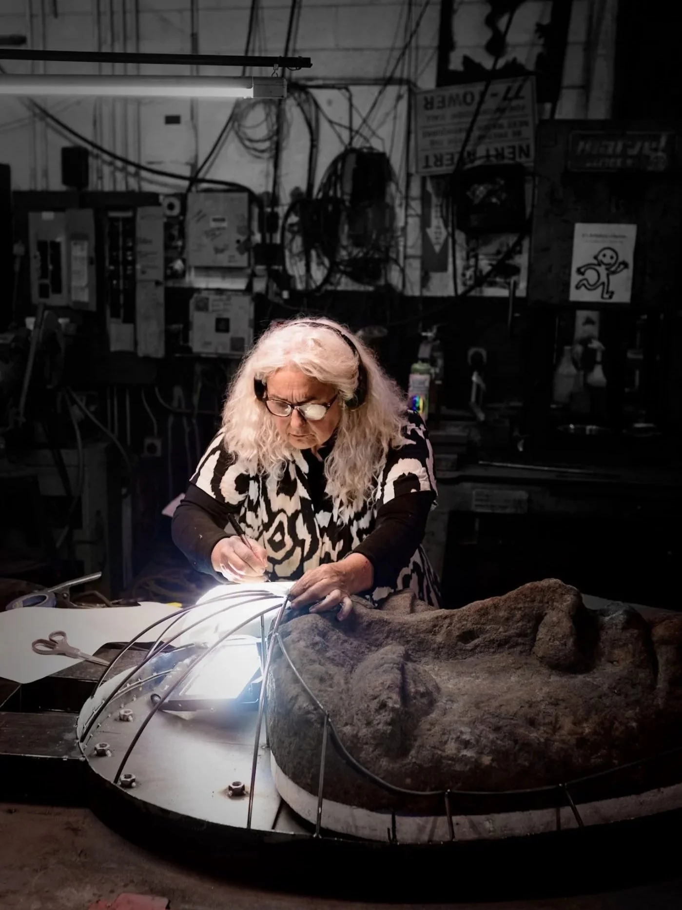 A woman with glasses and headphones working on a large face sculpture in a workshop, surrounded by tools and equipment.