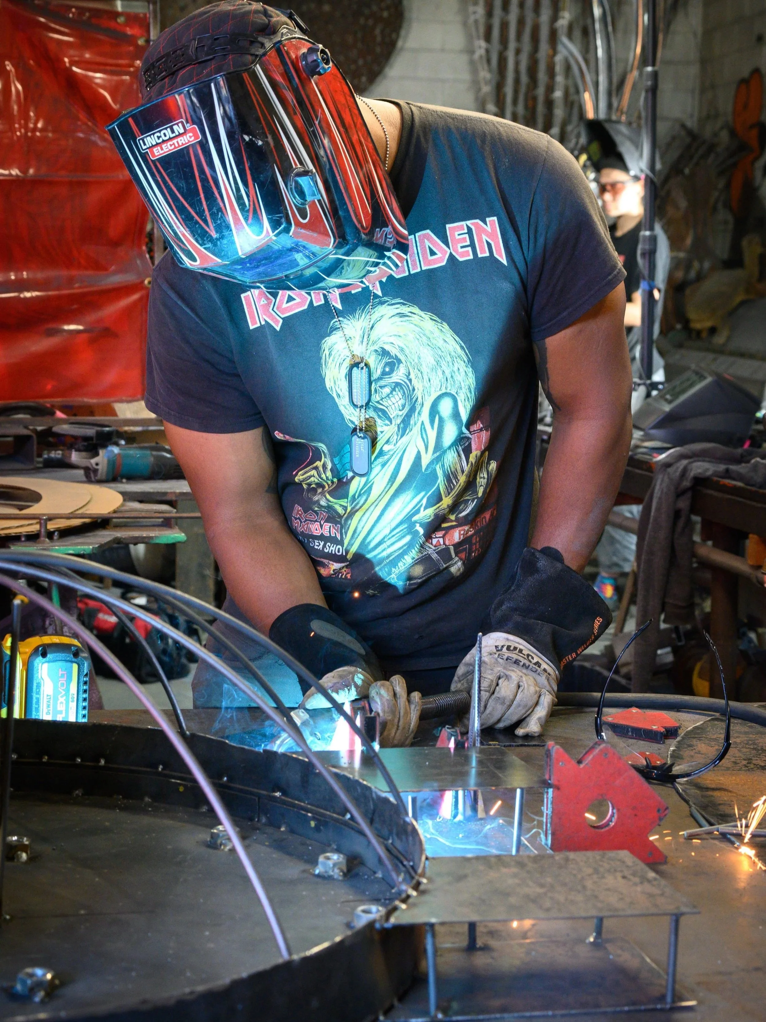 Artist Zulu Heru welding metal at a workshop, wearing a protective helmet and gloves, with equipment and tools around.