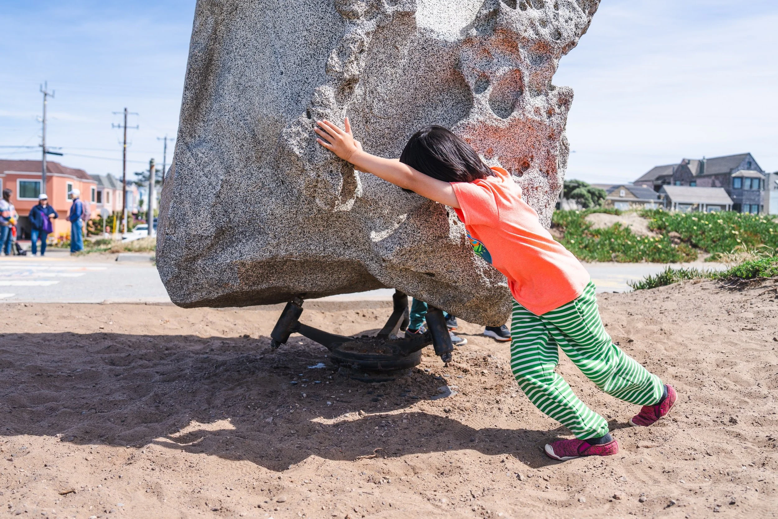 Child in orange shirt and green striped pants pushing a large boulder on a beach, with an urban background of houses and utility poles.