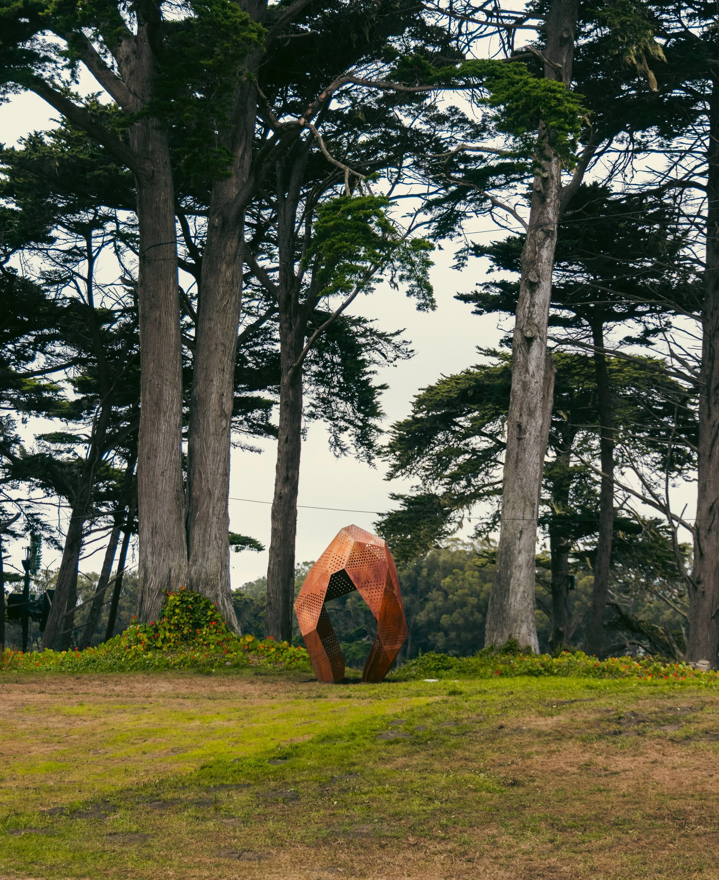 A park scene with large trees and green grass, featuring a modern, geometric, orange-brown sculpture in the center.