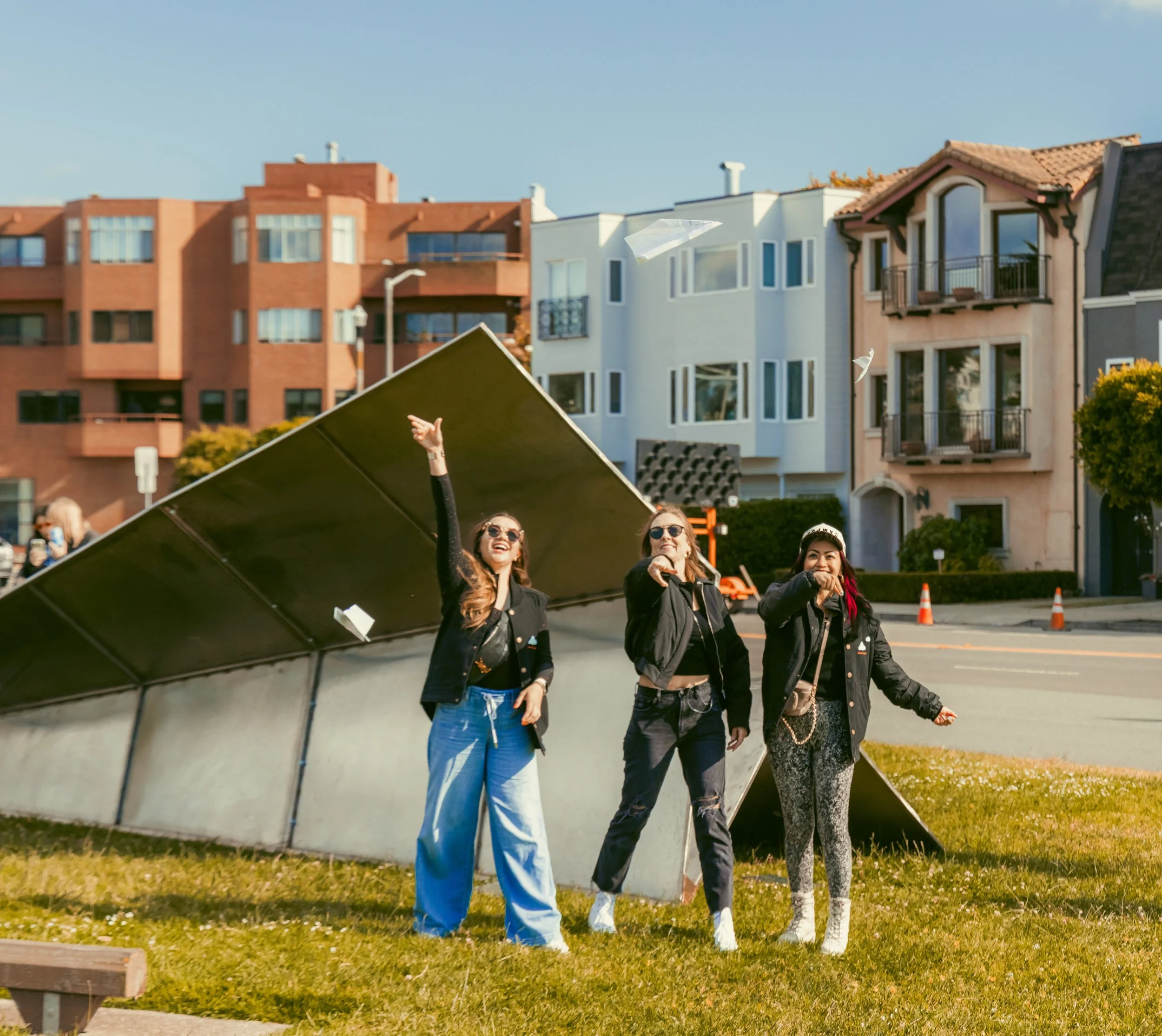 Three women in sunglasses smiling and raising their hands underneath an art installation resembling a tilted metallic roof, with residential buildings and trees in the background.