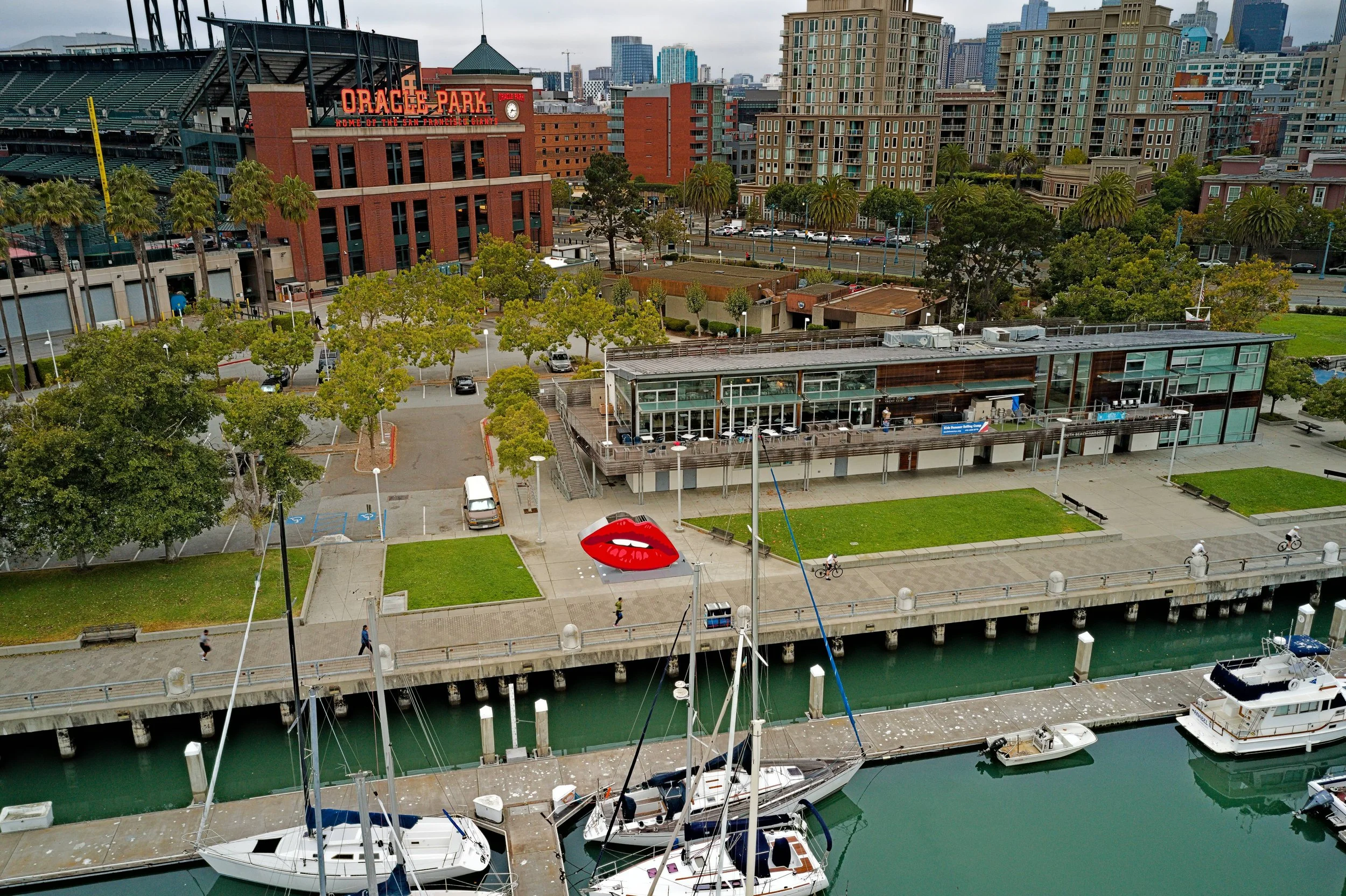 A cityscape featuring Oracle Park, a baseball stadium, with surrounding greenery, parking lots, modern buildings, and boats docked along a waterway in San Francisco, California.