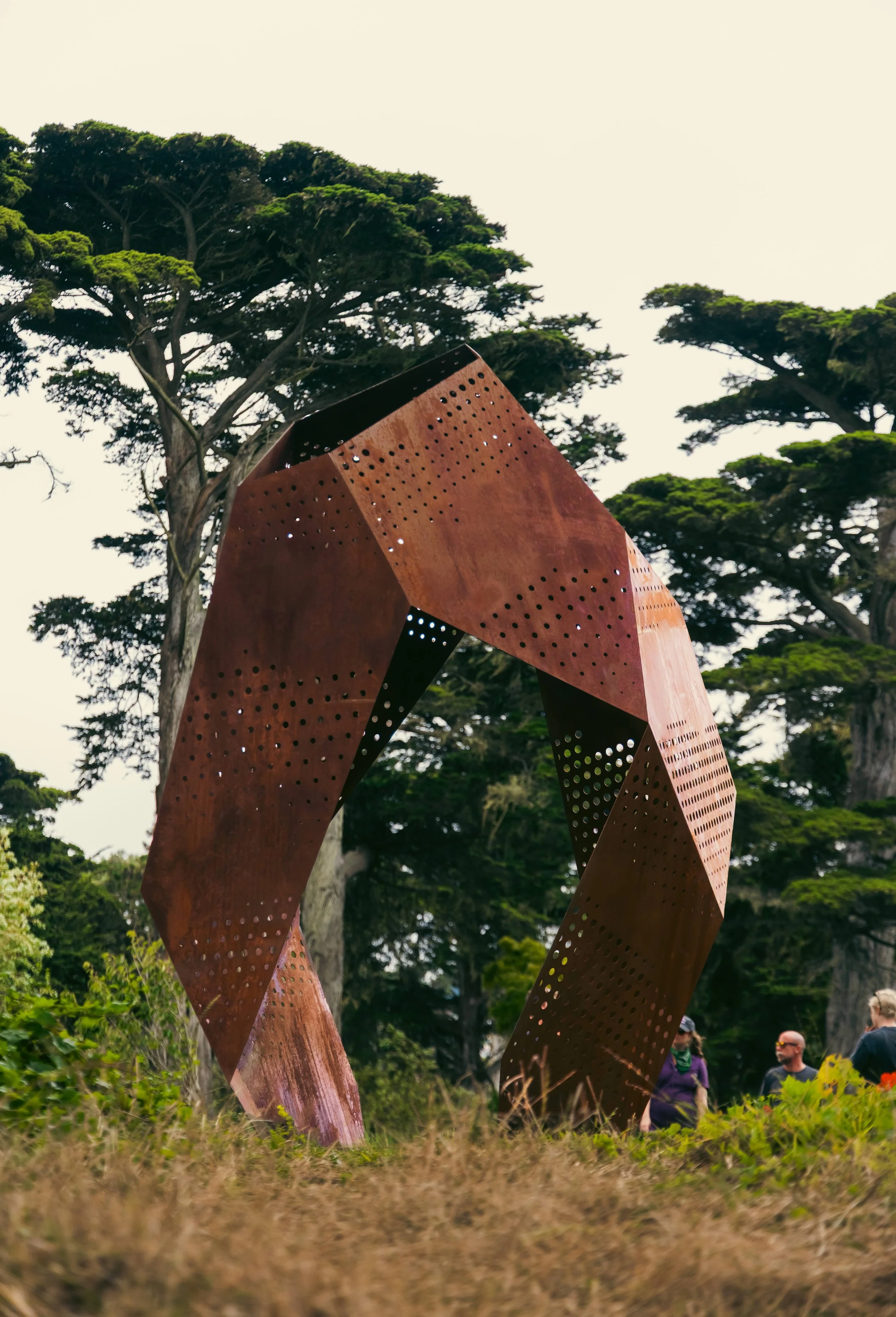 Large rust-colored metal sculpture with perforations, set against tall green trees and a cloudy sky, with a few people in the background.