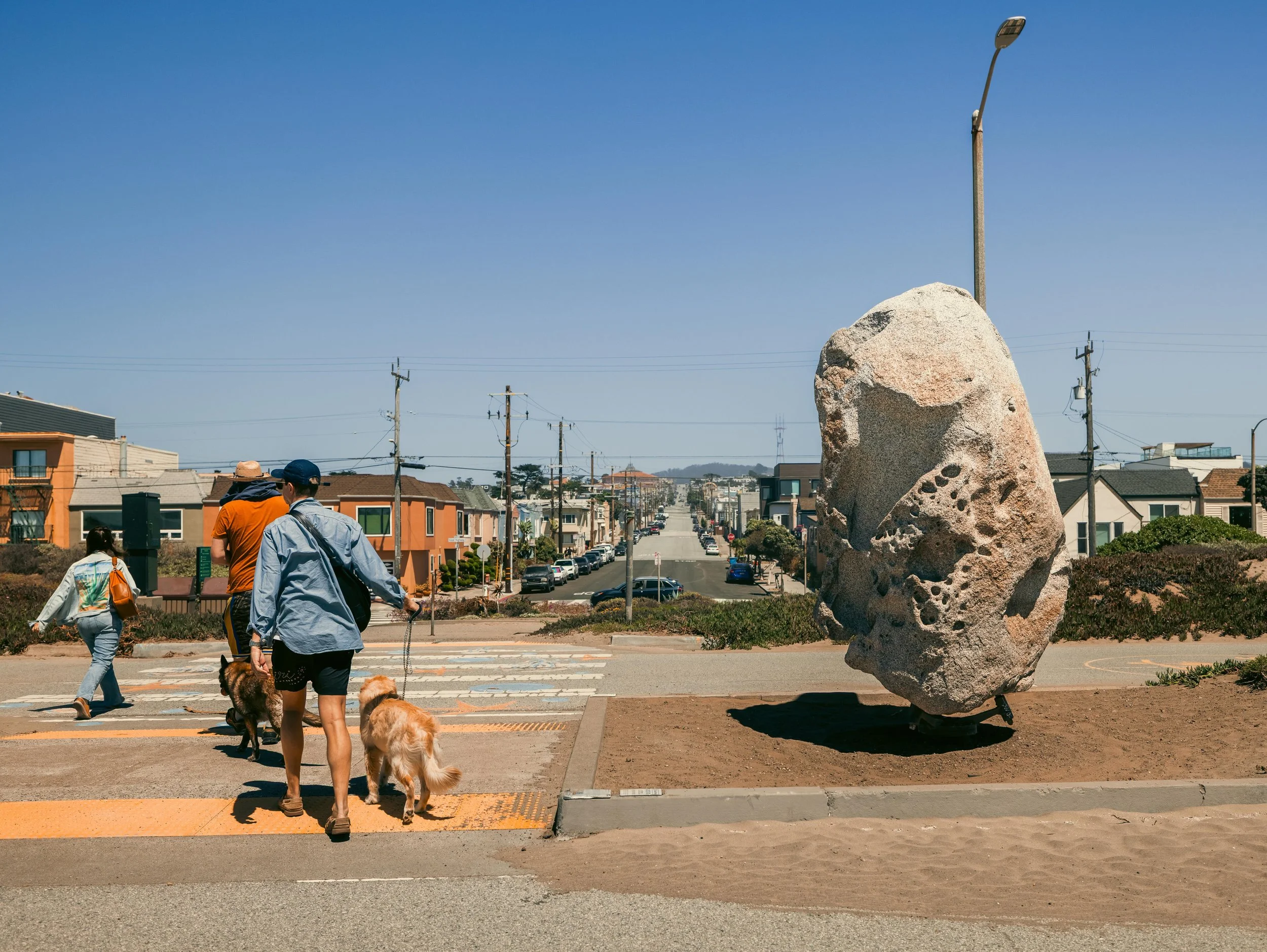 People walking with dogs across a crosswalk in an urban area with a large rock and a streetlamp in the background.