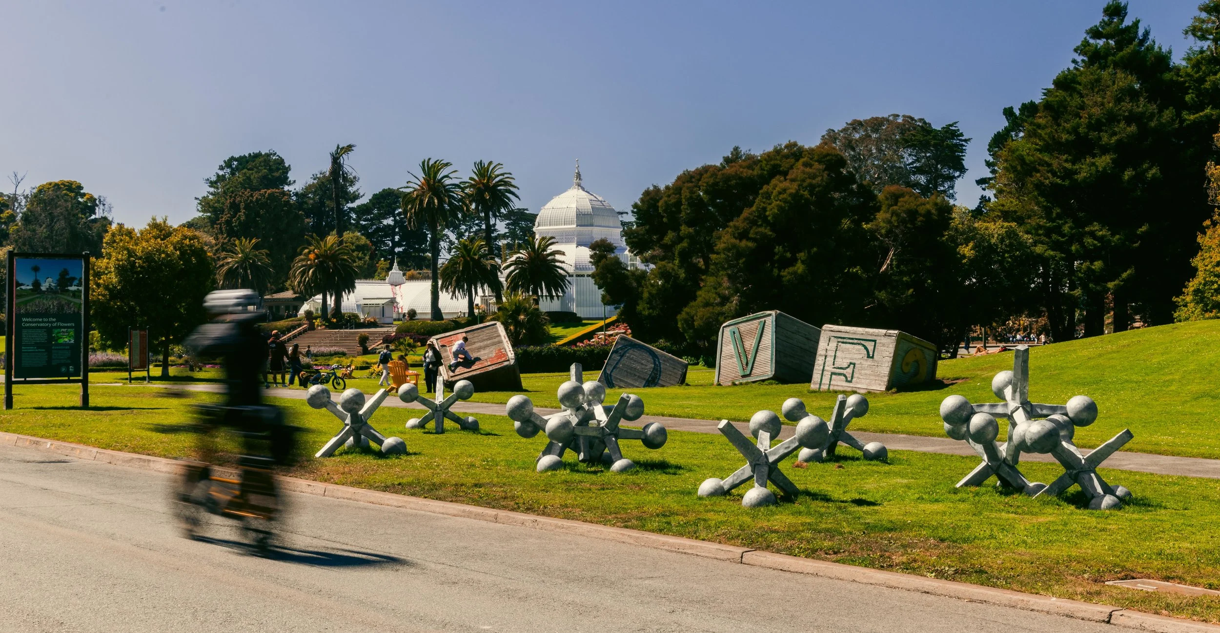 A park with large letter sculptures spelling 'LOVE' and molecular structures in the foreground, trees, and a white conservatory or greenhouse in the background. People are walking, sitting, and biking in the park.
