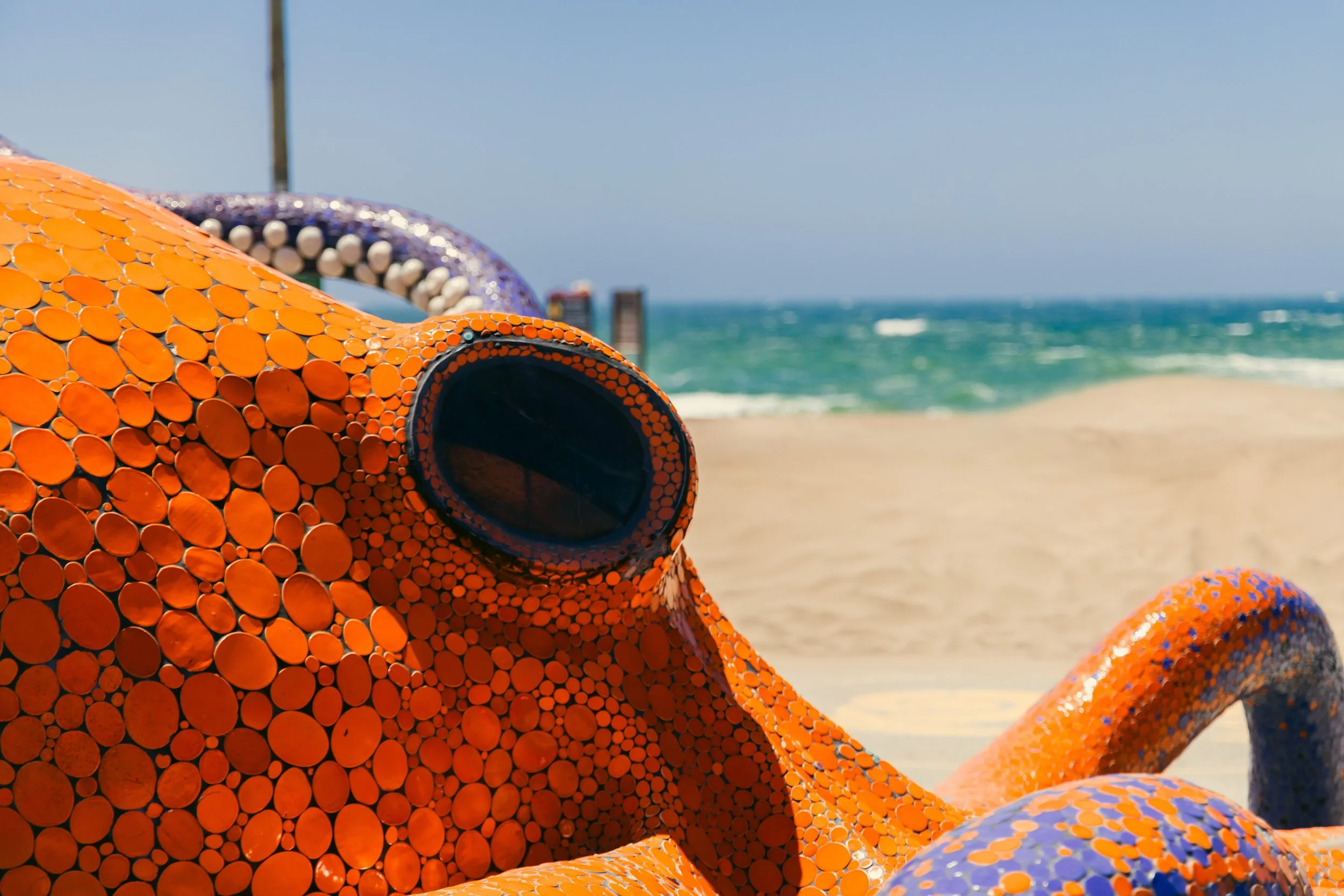 A close-up of Octavius, a mosaic octapus sculpture, made of orange, purple, and white tiles, on a beach with the ocean and clear sky in the background.