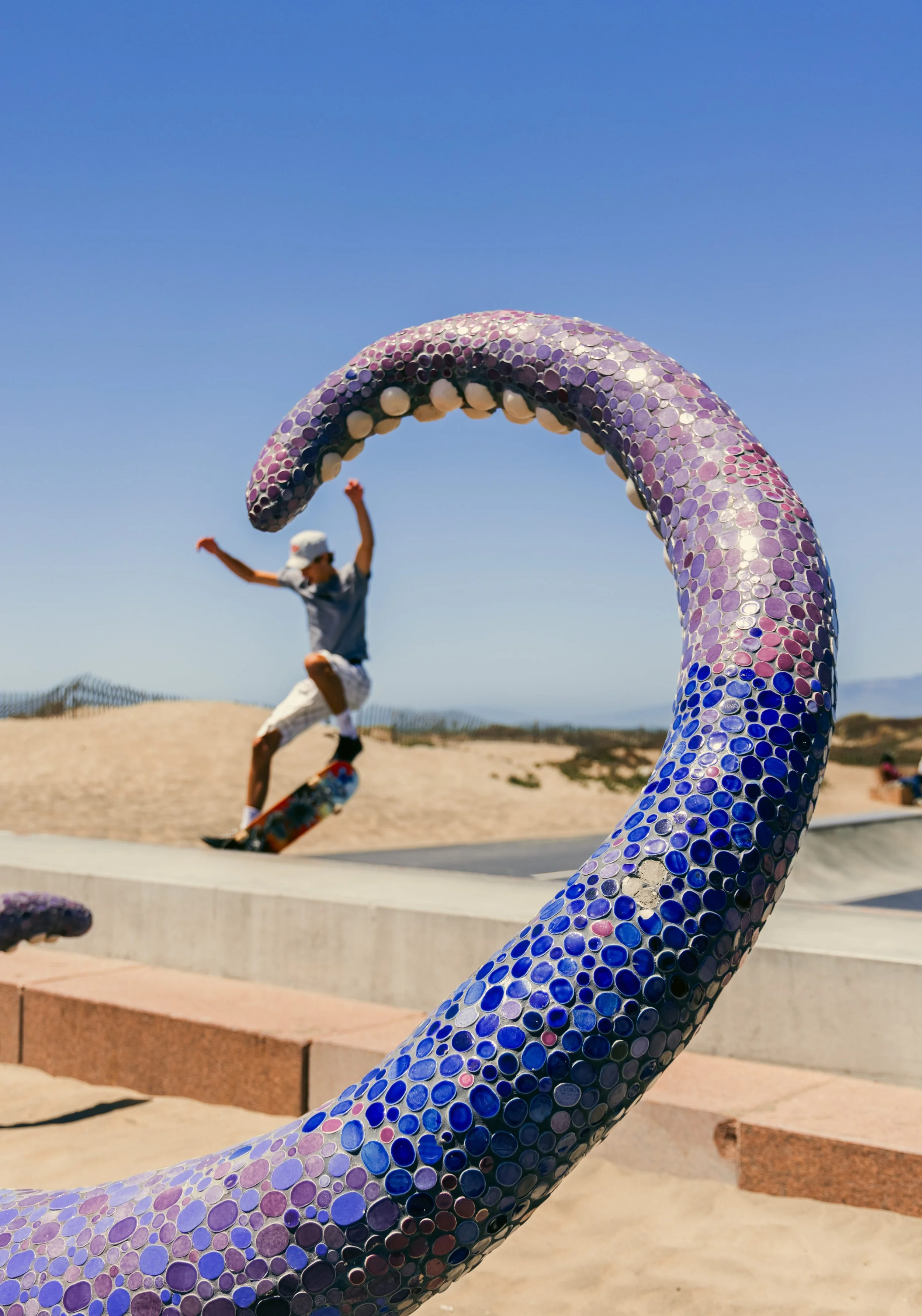 A person skateboarding in the background with a large, colorful, mosaic sculpture of a snake in the foreground at a beach side park.