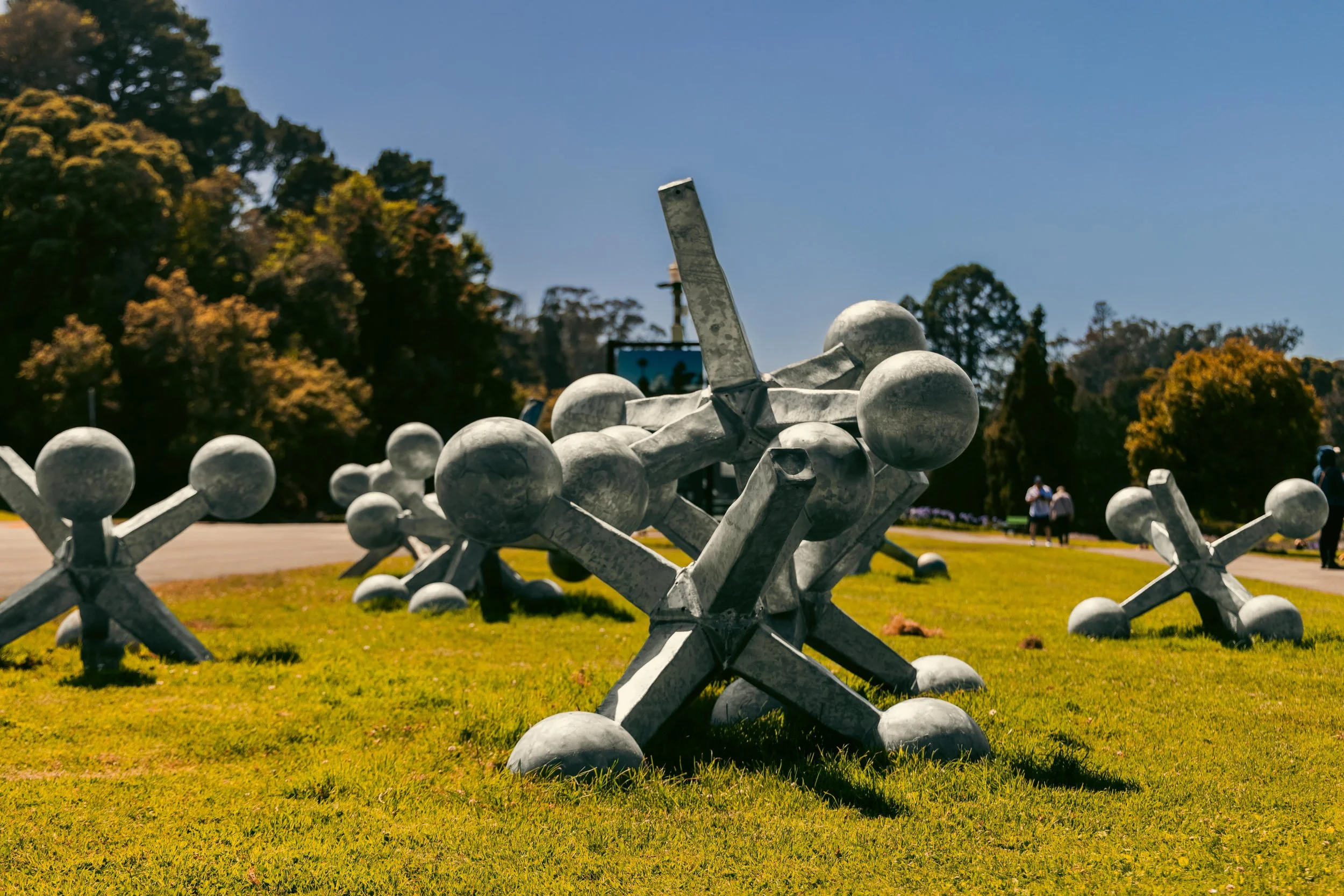 Sculptural art installation made of interconnected metal spheres and rods, situated on a grassy area in a park with trees and blue sky in the background.