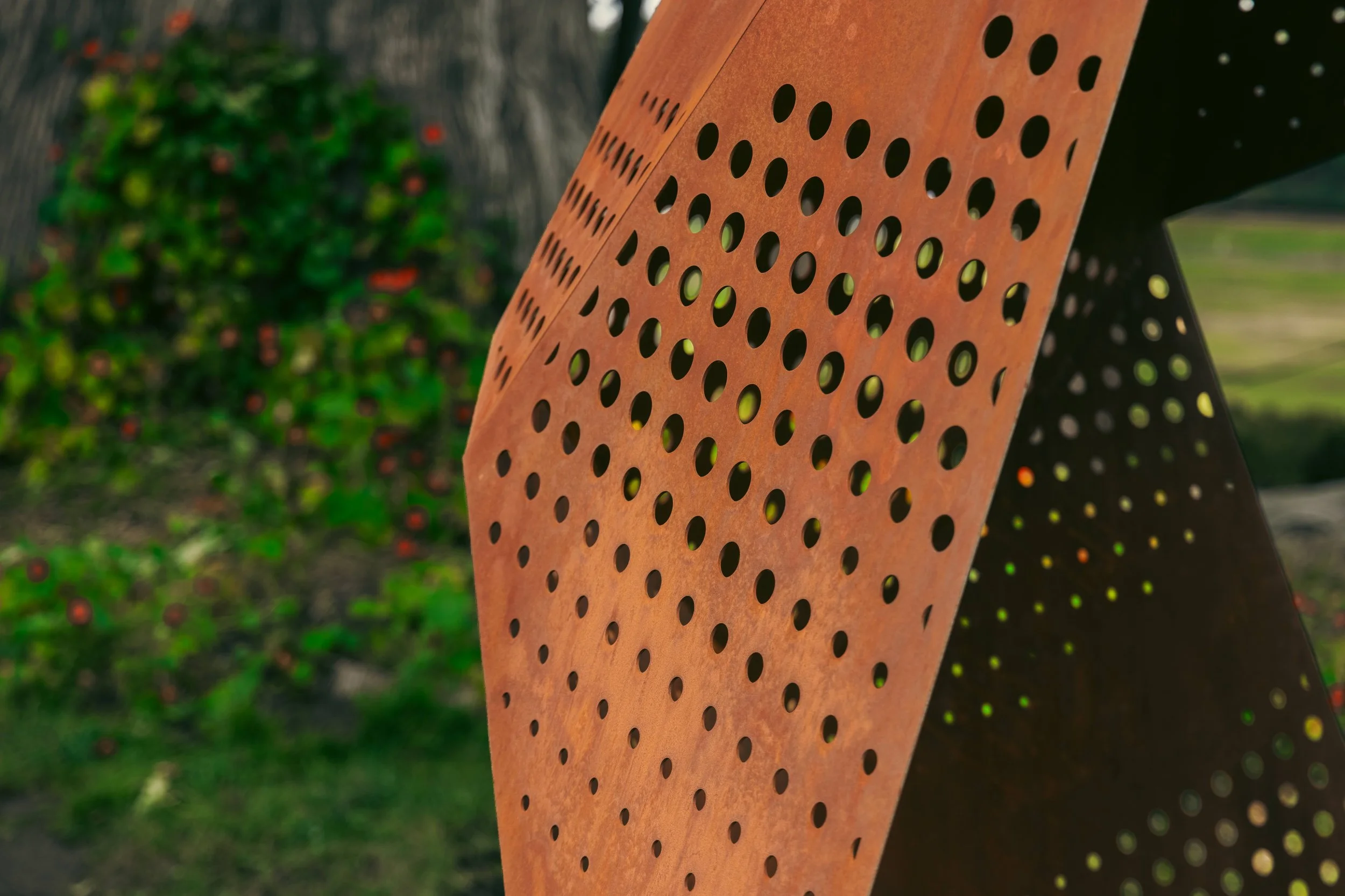Close-up of a rust-colored metal sculpture with multiple circular holes arranged in a pattern, outdoors with blurred greenery in the background.