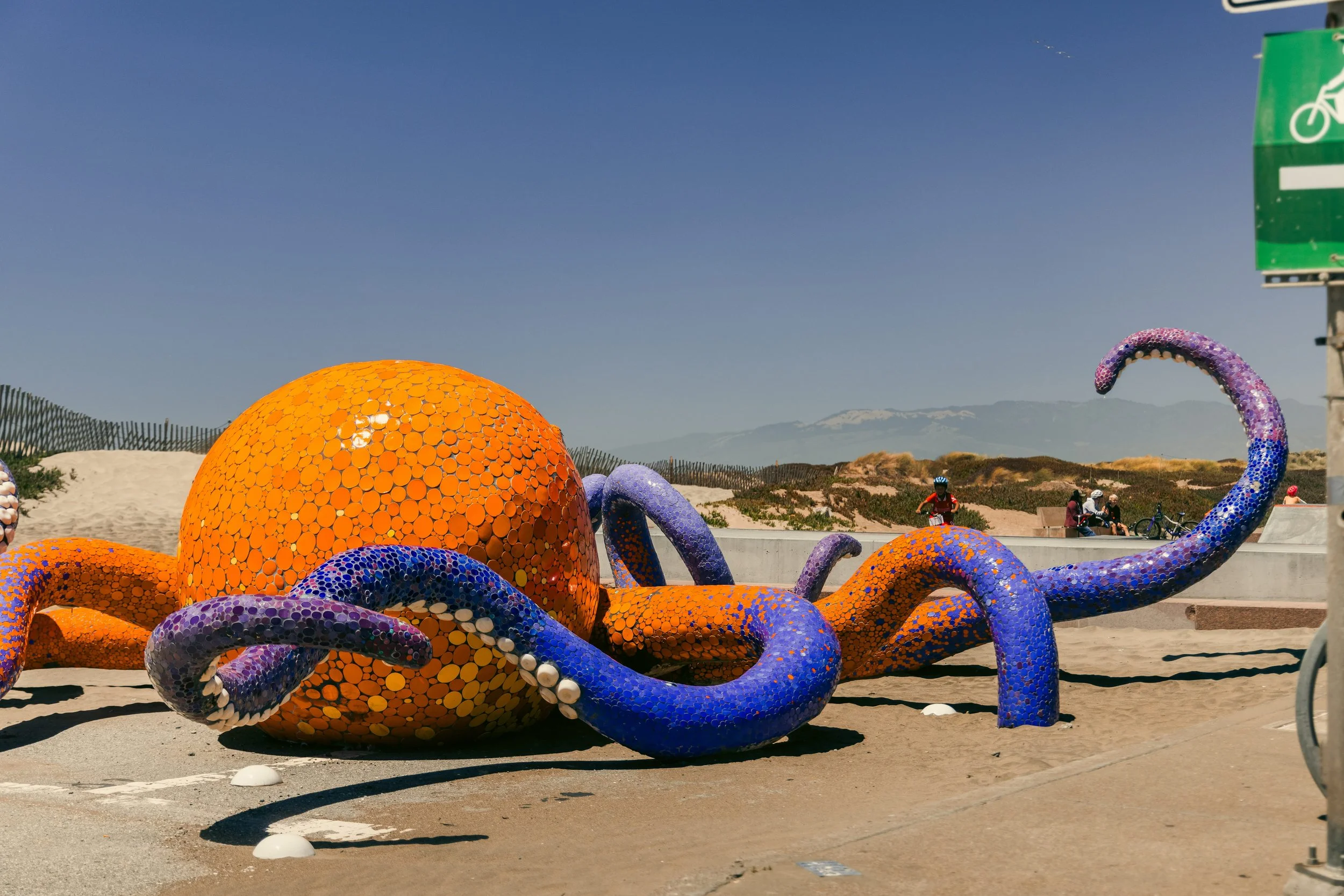 Colorful octopus sculpture made of mosaic tiles at a beachside park, with people in the background and green bike path sign to the right, mountains in the distance under a clear blue sky.
