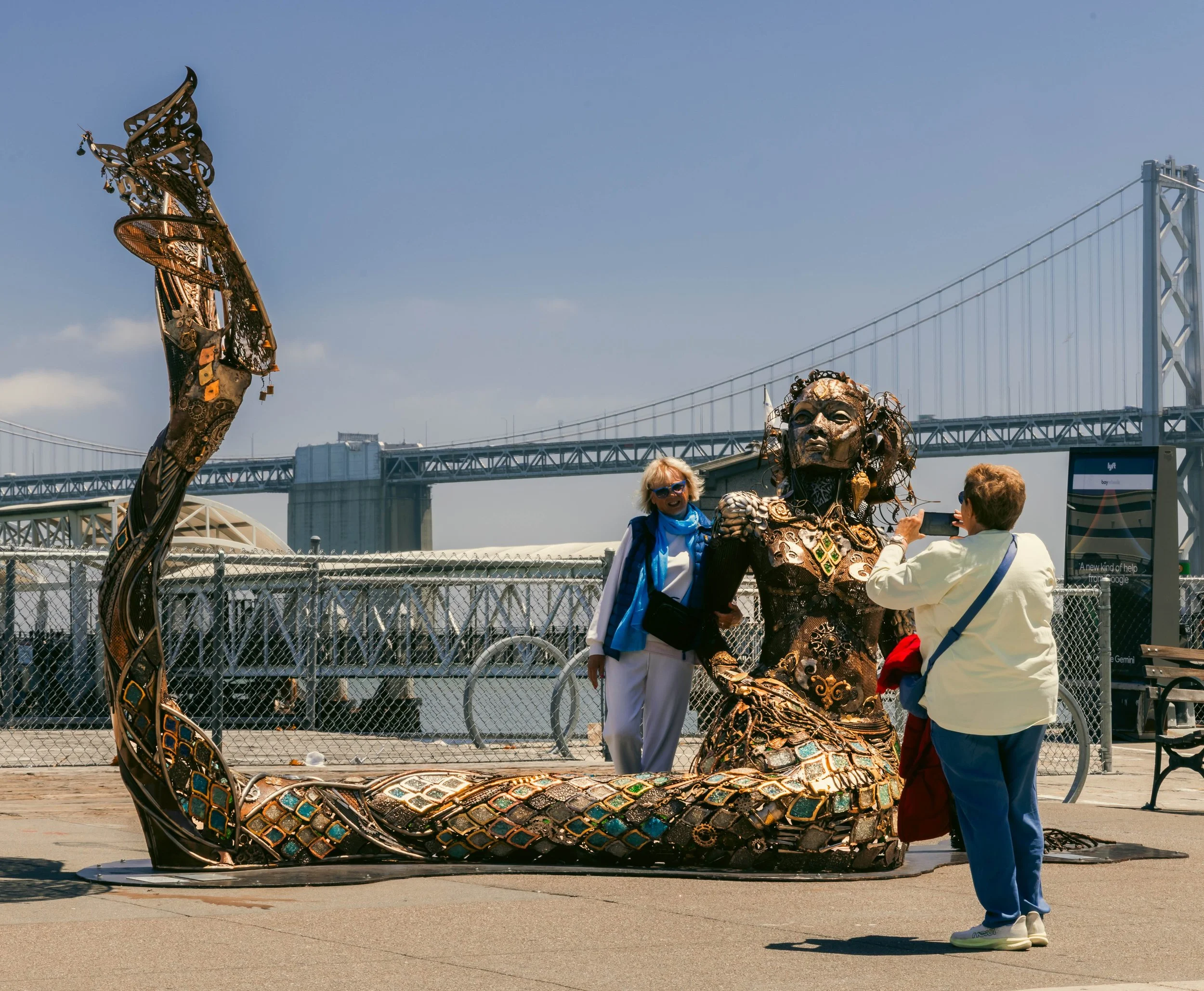Two women in conversation, with a city bridge in the background standing in front of a sculpture called Coralee by artists Dana Albany.