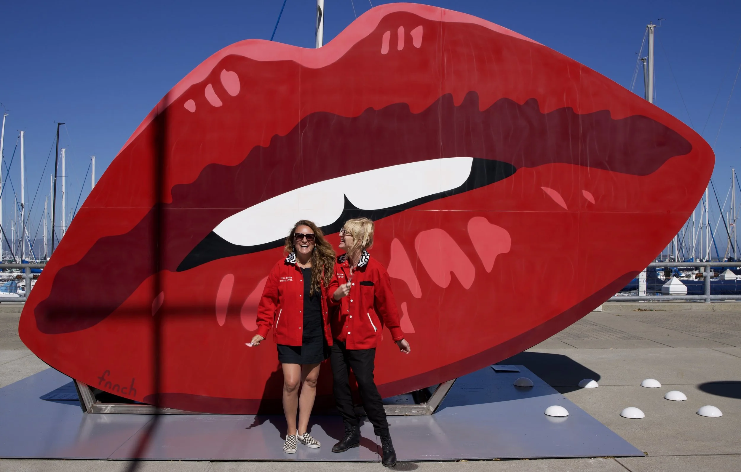 Two women standing in front of a large red art installation of lips with an open mouth, at a marina with sailboats in the background. One woman is wearing sunglasses, a red jacket, and a black skirt, while the other has blonde hair, glasses, and is dressed in black and red.