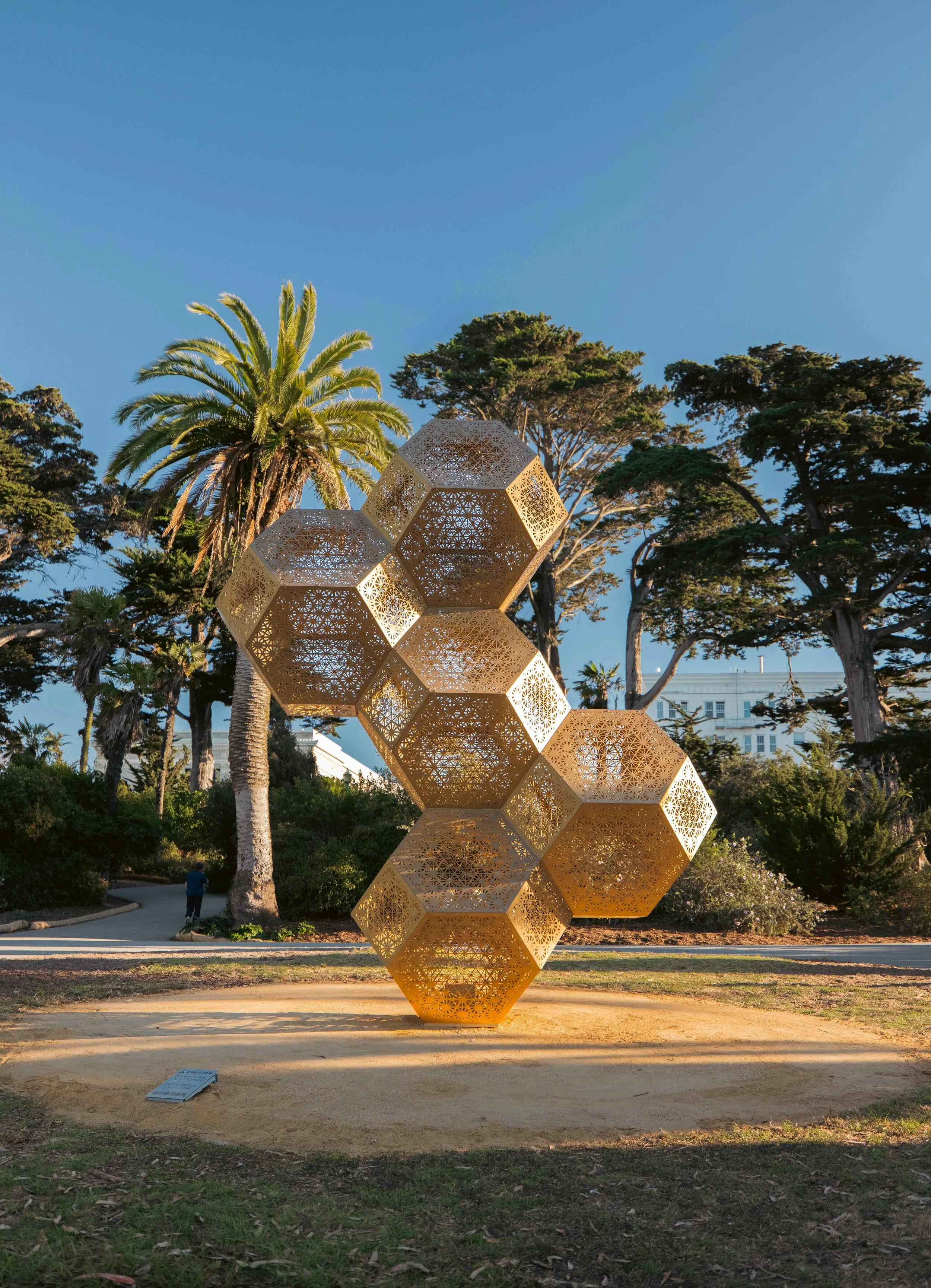 Large geometric sculpture made of interconnected golden hollow polyhedrons installed outdoors, with a person standing beside it for scale and a cityscape in the background. The text "COMING SOON" overlays the image.