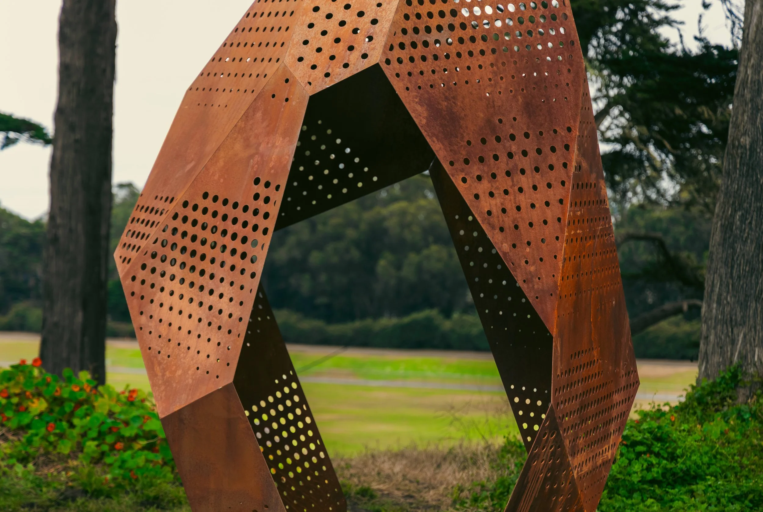 Rust-colored metal sculpture with perforations, shaped in a looping curvilinear form, set outdoors with trees and a grassy area in the background.