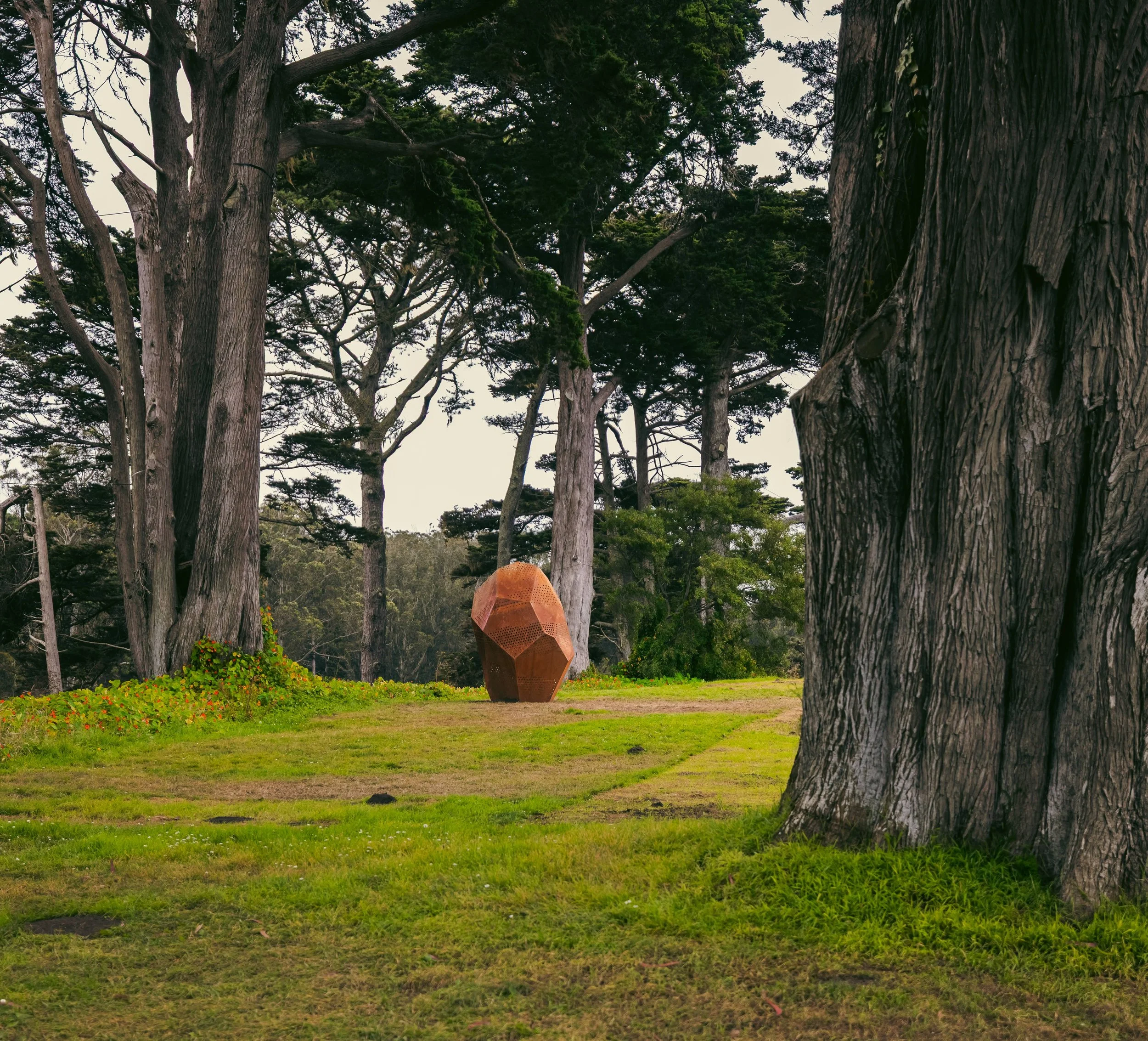 Trees in a forest with a grassy clearing, a person with an orange umbrella walking, and a geometric sculpture in the background.