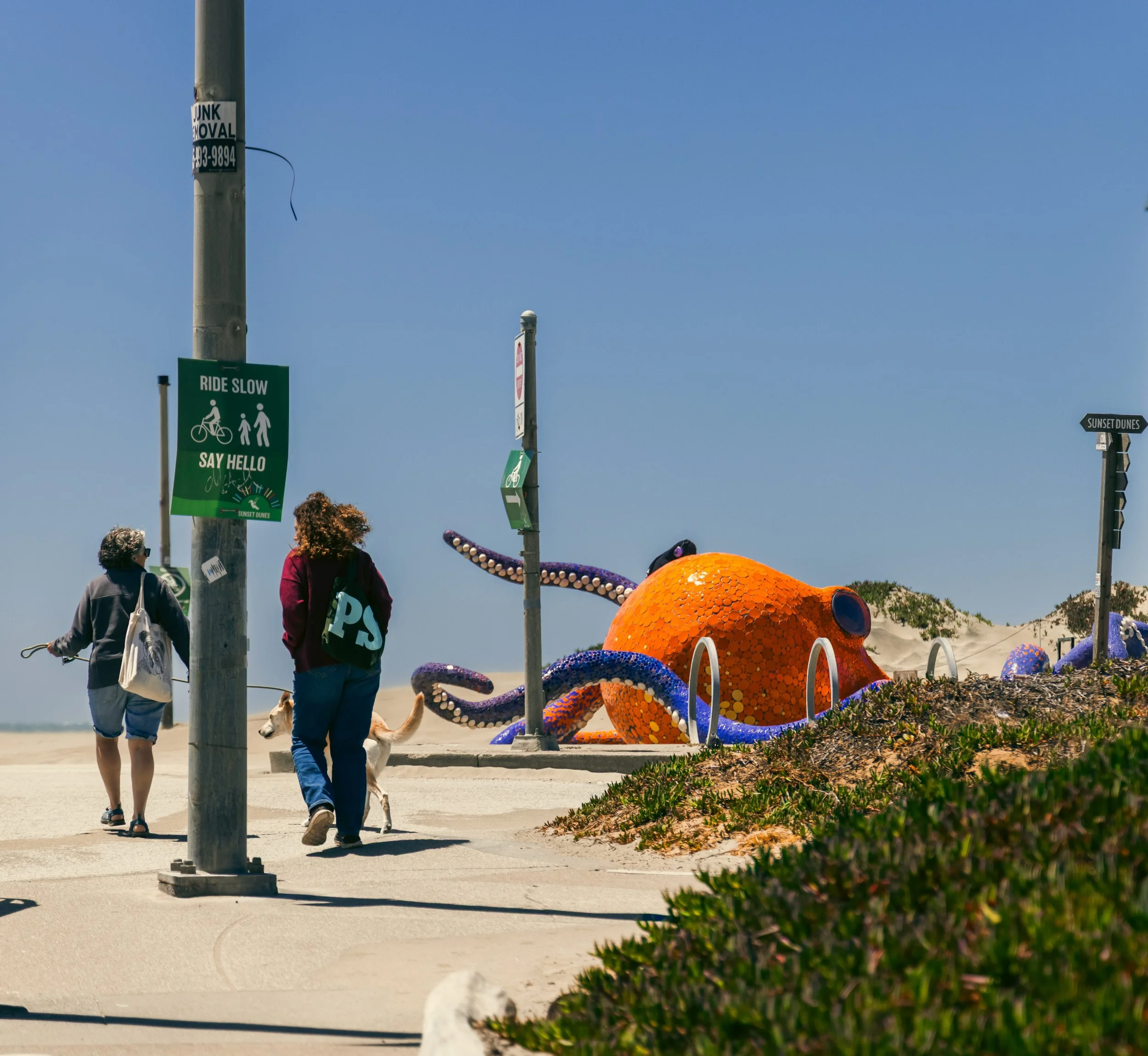 People walking along a sidewalk near beach-themed sculptures and signs, with clear blue sky overhead.