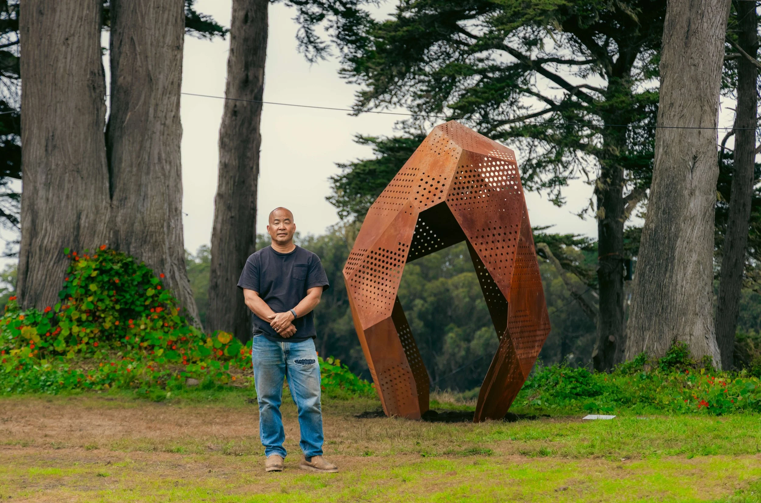Artist Miki Iwasaki stands outdoors in front of large trees and a modern, perforated metal sculpture. There is a bush with red flowers on the left side of the image.