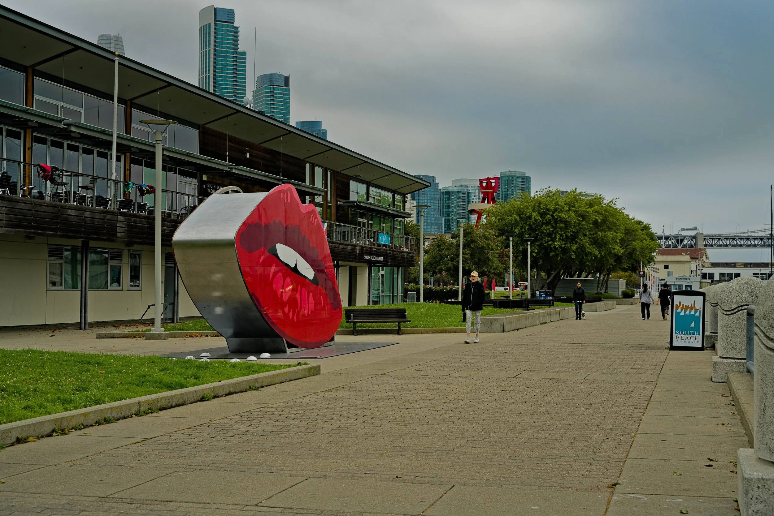 Outdoor scene at South Beach Harbor with a large red art installation of lips, a modern building with balconies, and tall skyscrapers in the background, overcast sky, people walking on paved path, some greenery and trees.