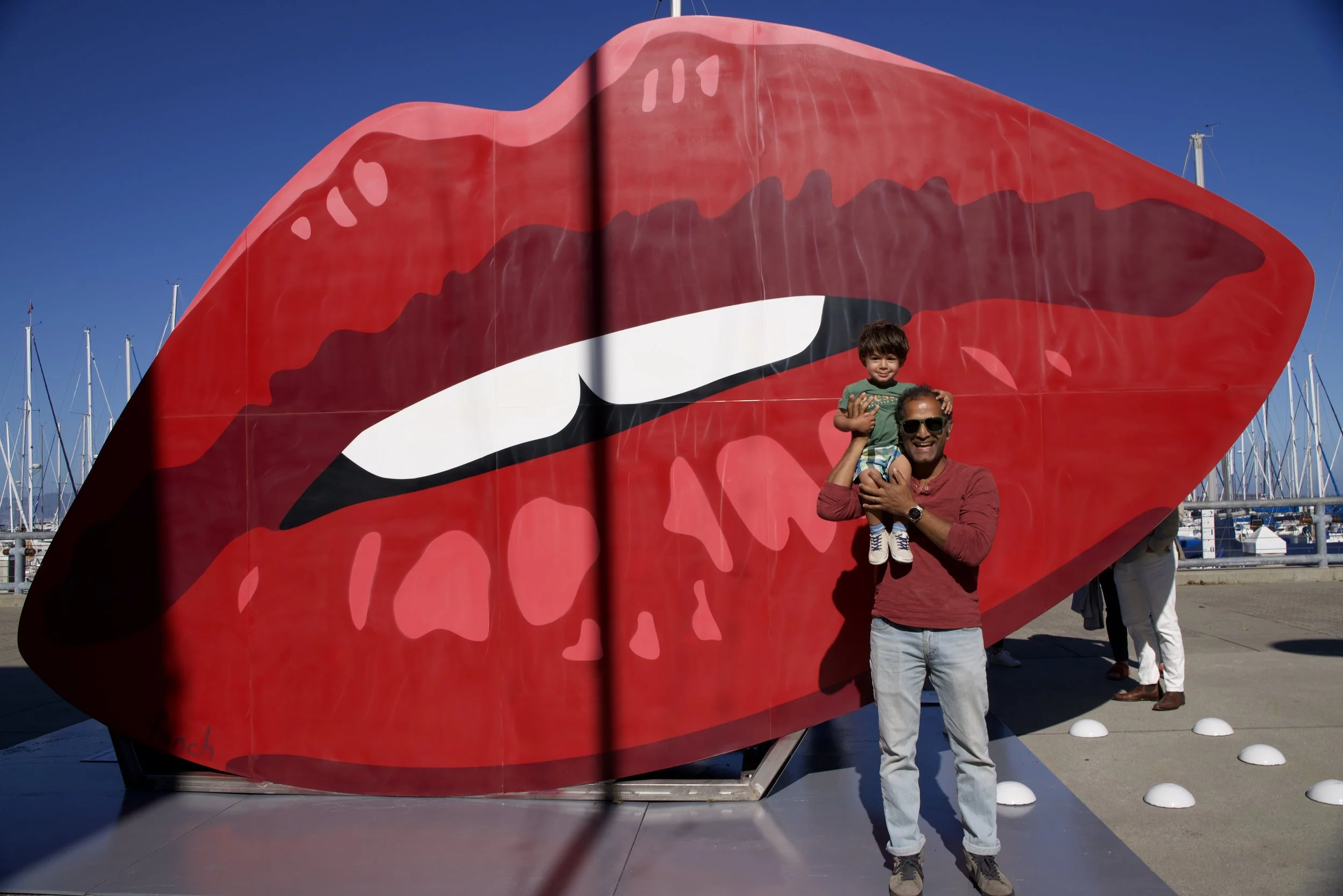 A man is holding a young boy on his shoulders in front of a large, colorful lips mural. The mural features open lips with red lips, black outline, and white teeth, set against a blue sky with sailboat masts in the background.